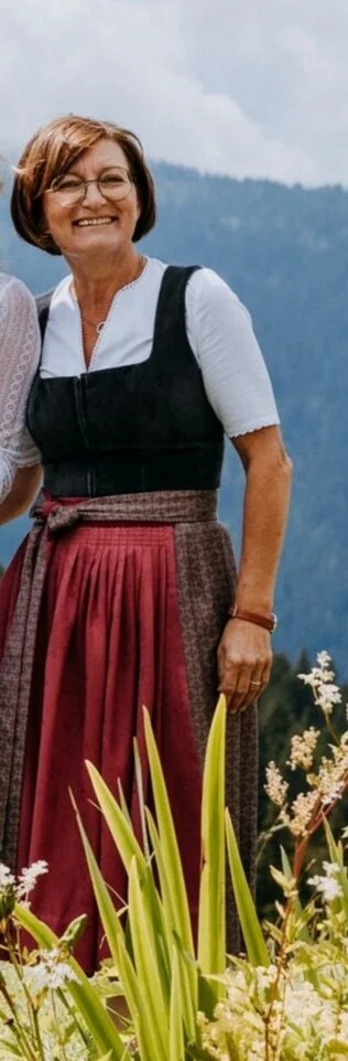 Frau in traditioneller Kleidung vor einer Berglandschaft mit Blumen im Vordergrund.