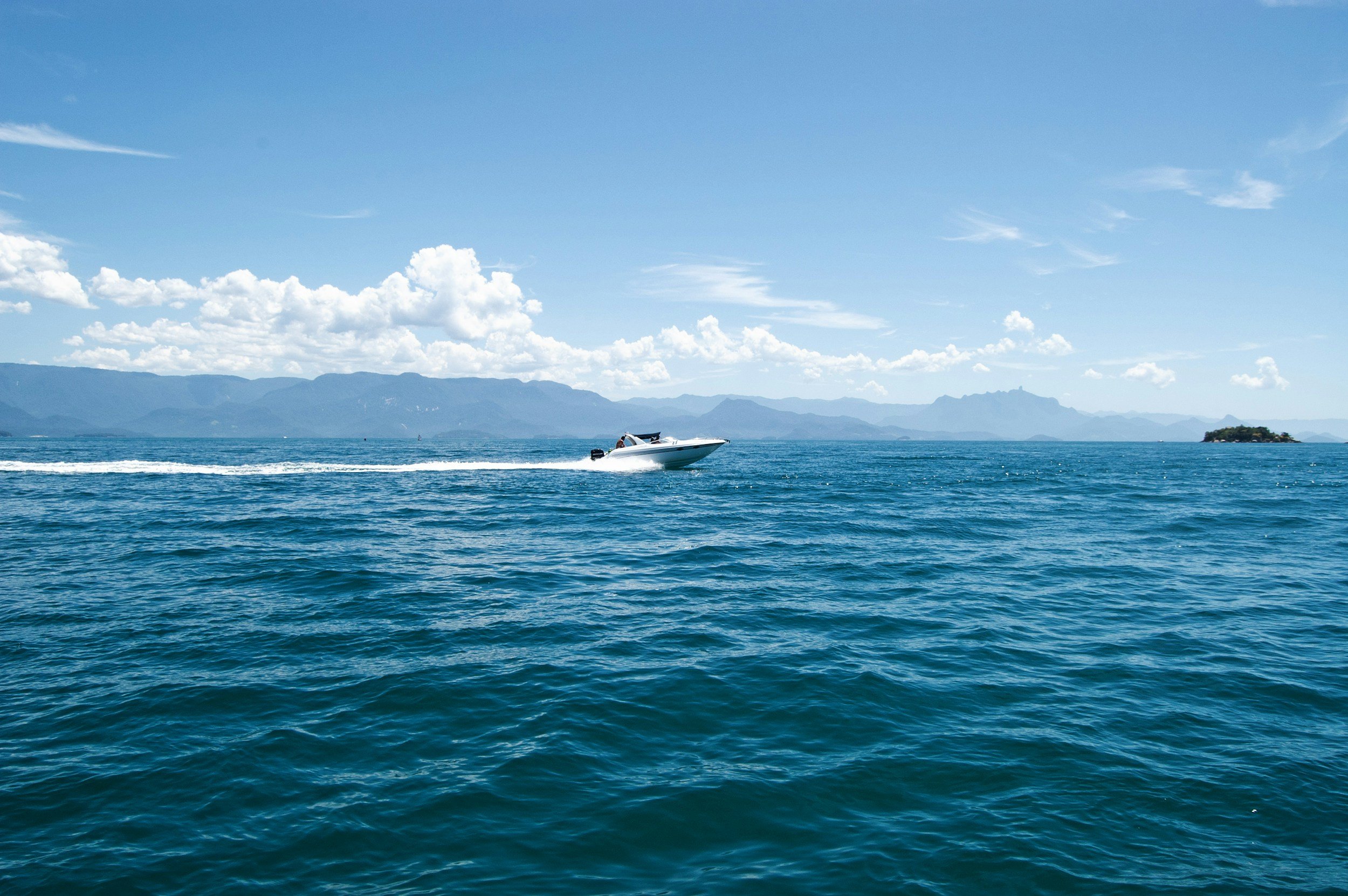 Ein weißes Motorboot auf dem blauen Meer mit einer Insel und Berg im Hintergrund, unter bewölktem Himmel.