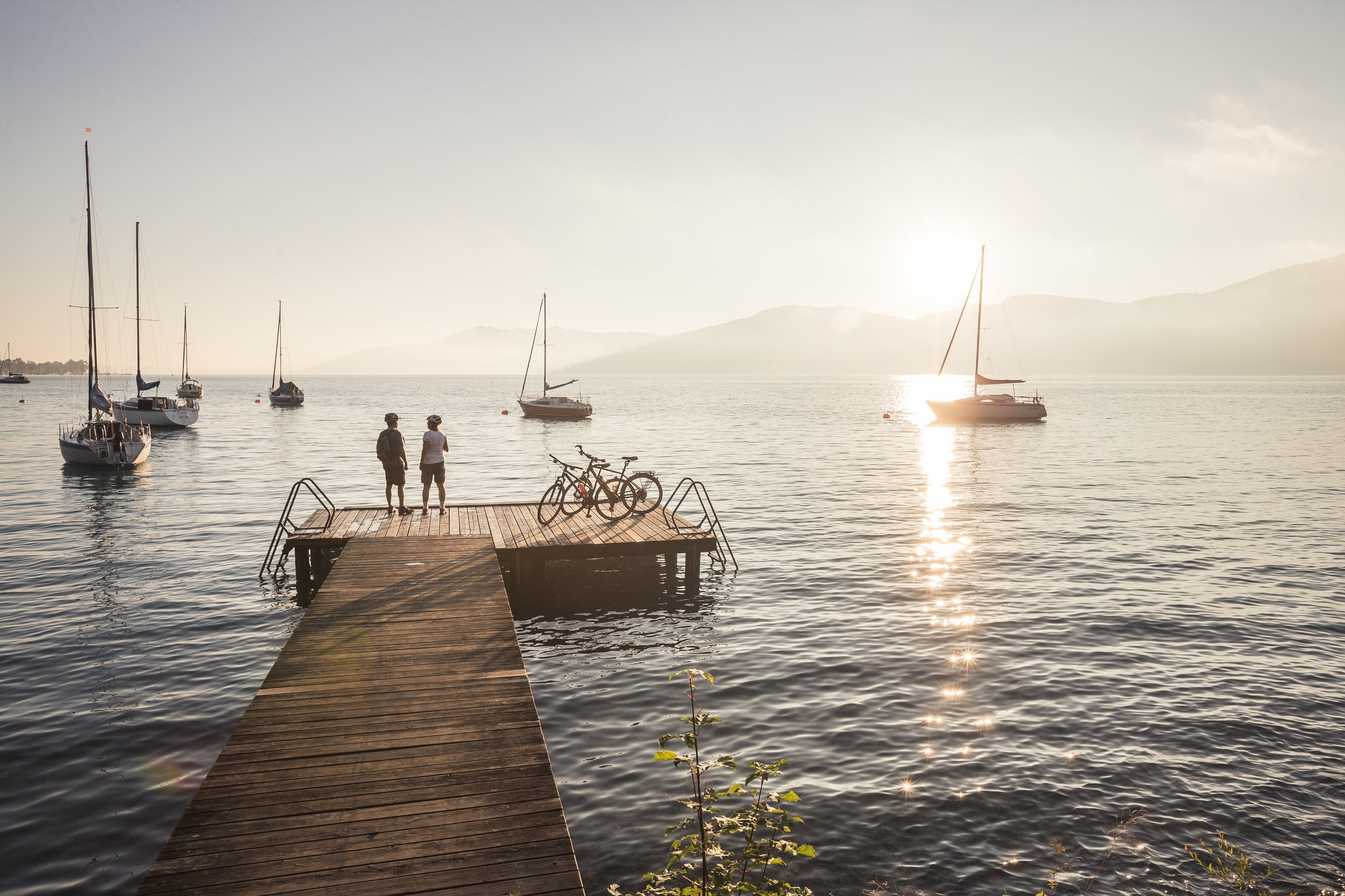 Zwei Personen stehen auf einem Steg am See, betrachten die Boote und den Sonnenuntergang.