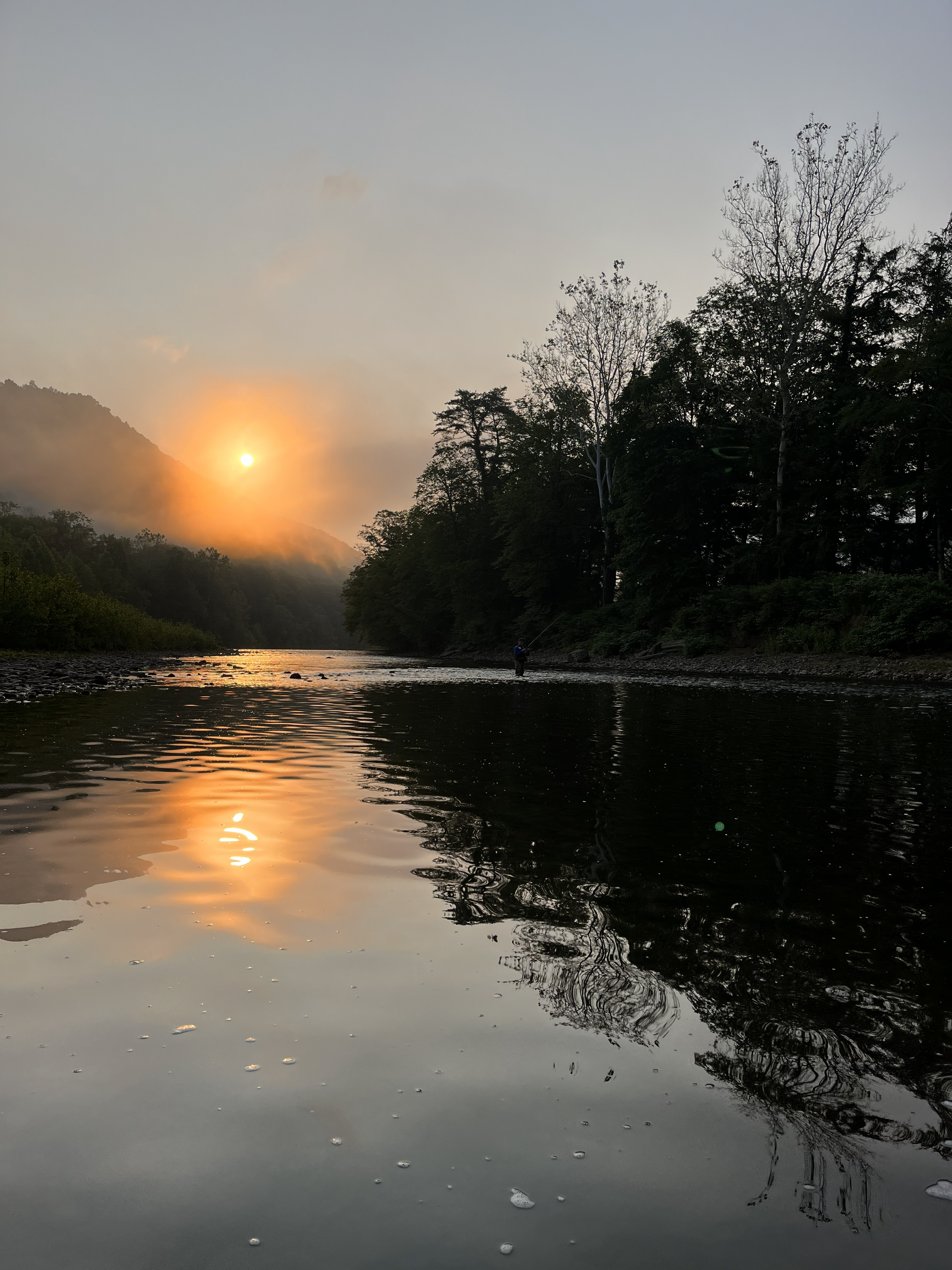 Sunset over a river surrounded by trees, with a person fishing on the riverbank and the sun's reflection on the water.