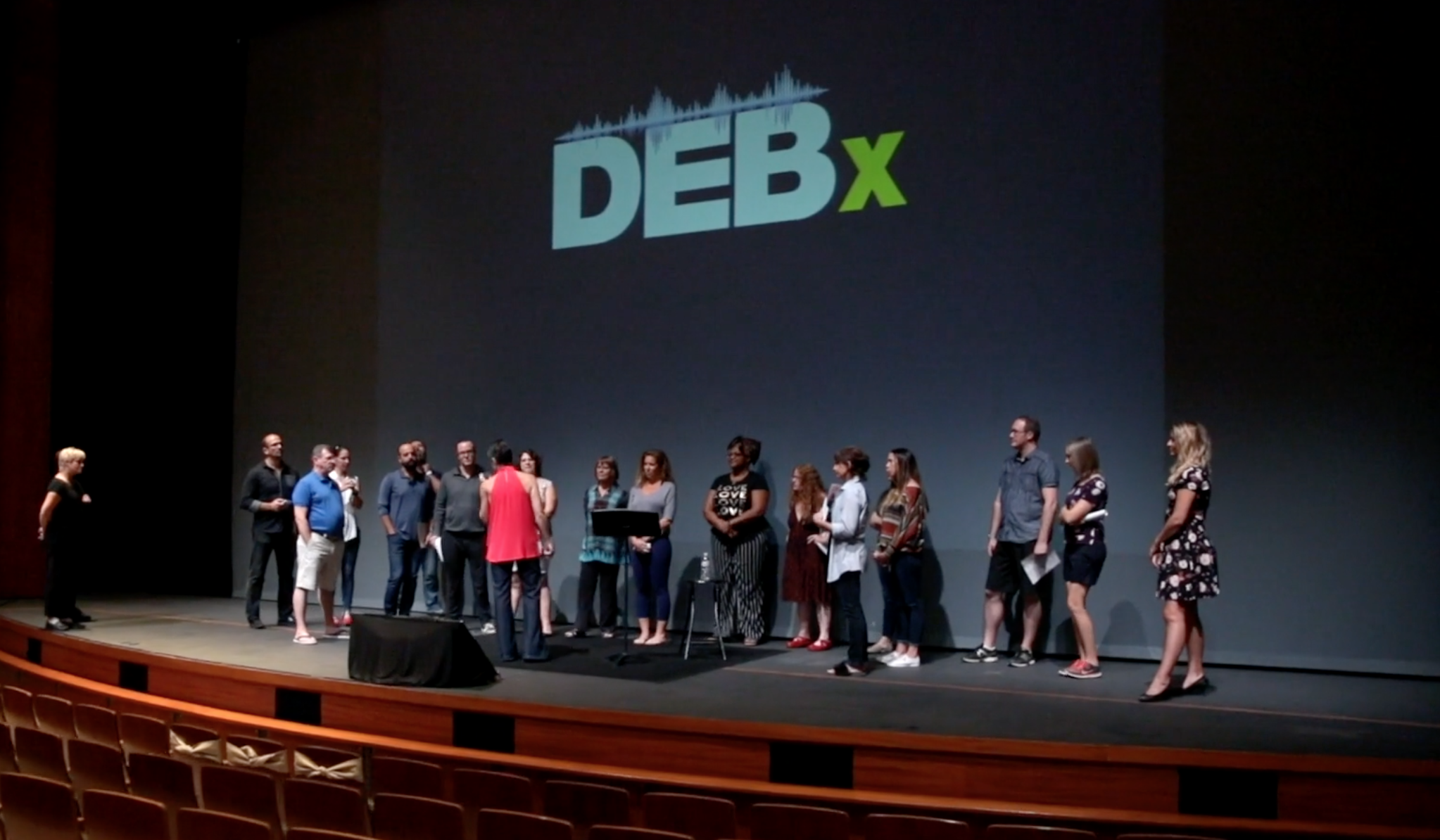 Group of people standing on stage in front of a large screen displaying the word 'DEB' with a green 'X' at an event or conference.