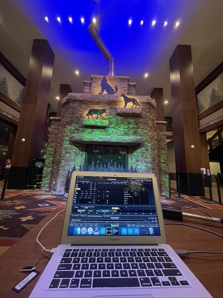 Interior view of a hotel lobby with a rock waterfall feature decorated with animal cutouts, illuminated with colorful lighting, and a DJ laptop setup in the foreground.