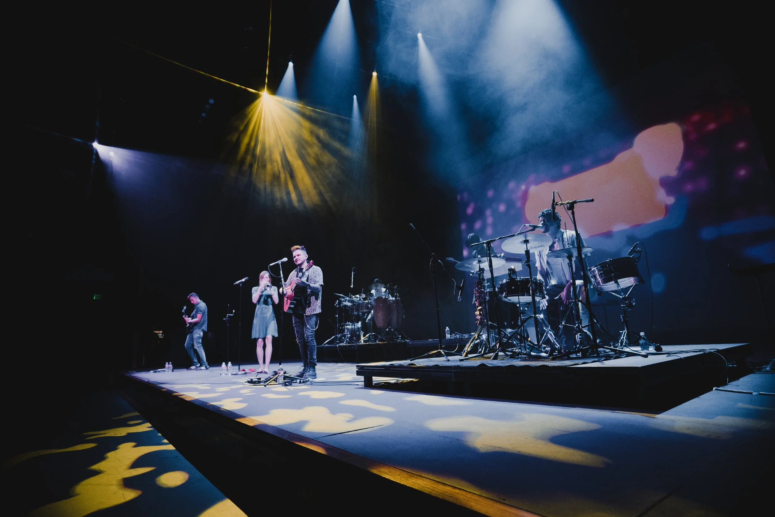 A band of multiple musicians performing on stage with bright stage lights and a large screen displaying a pink background with a pink flower.
