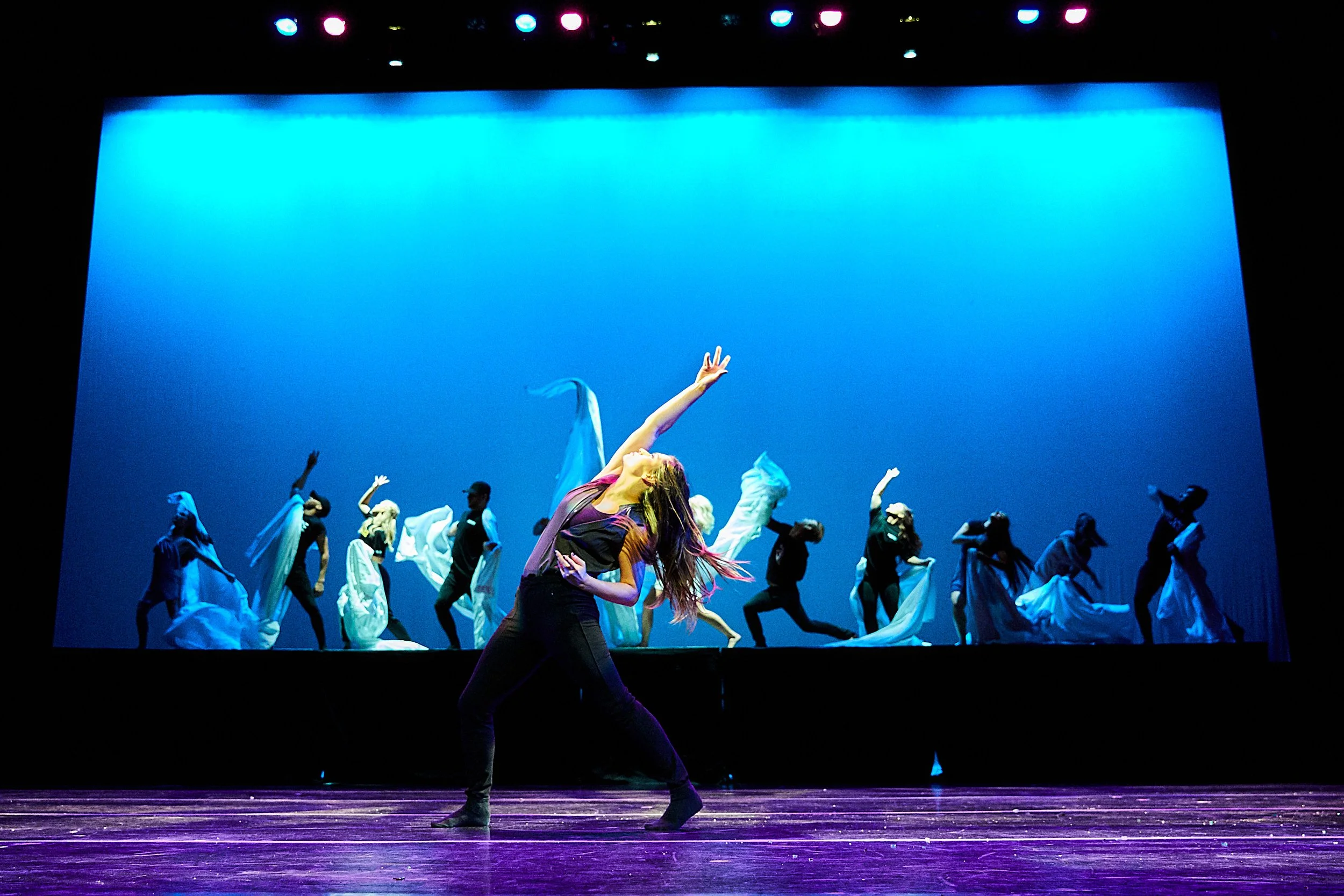 Group of dancers performing on a stage with a blue background, one woman in the foreground with long hair and dressed in black, reaching upward with one arm.
