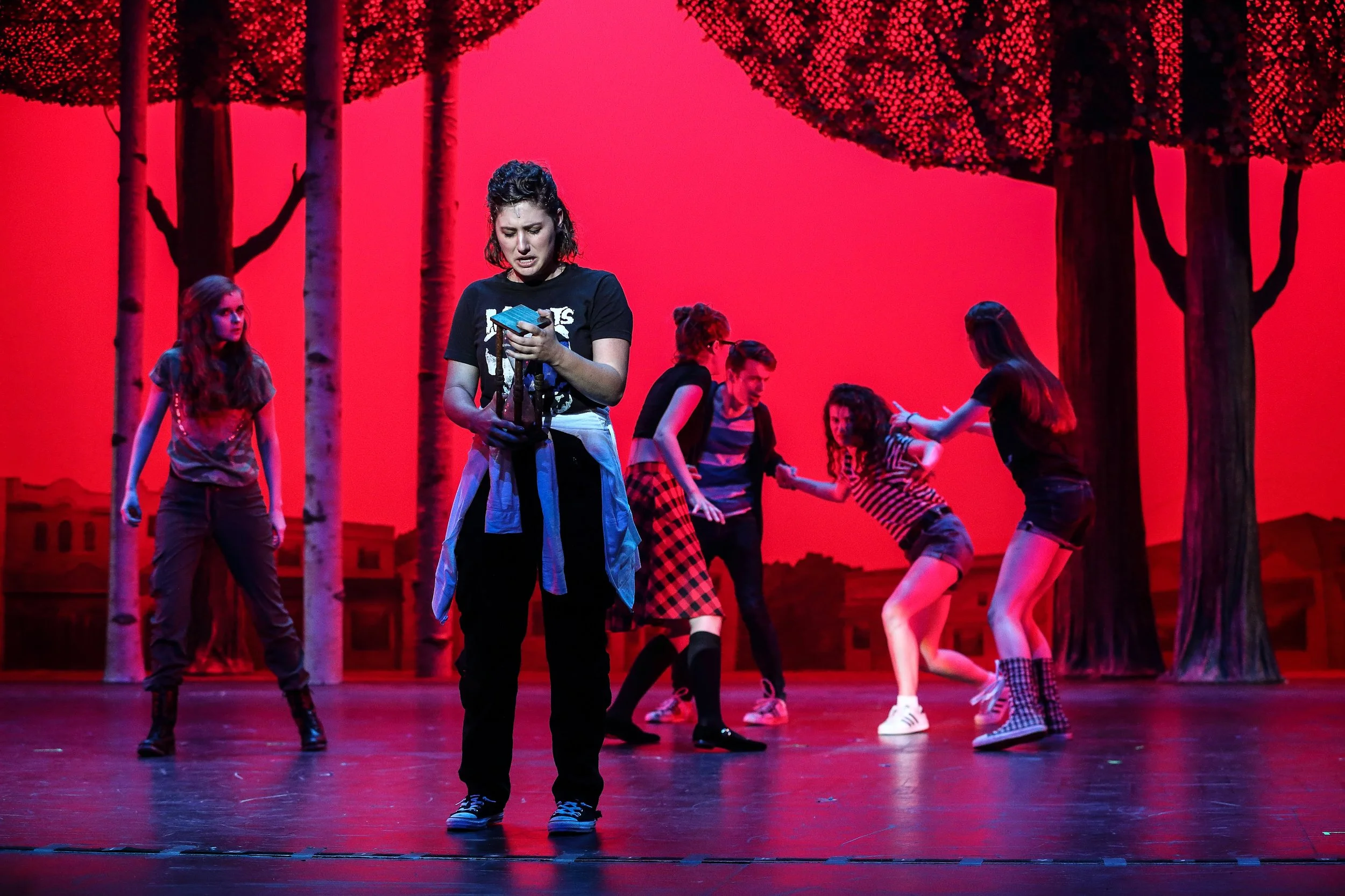 A group of young women performing on stage with a vibrant red background, surrounded by stylized trees, with one woman at the forefront holding a phone and others engaged in dance.