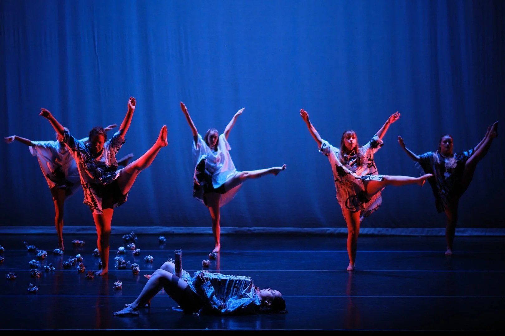 Five dancers perform on stage with a dark blue curtain backdrop, dressed in patterned pajamas, one dancer lying on the floor while the others balance on one foot with arms extended.