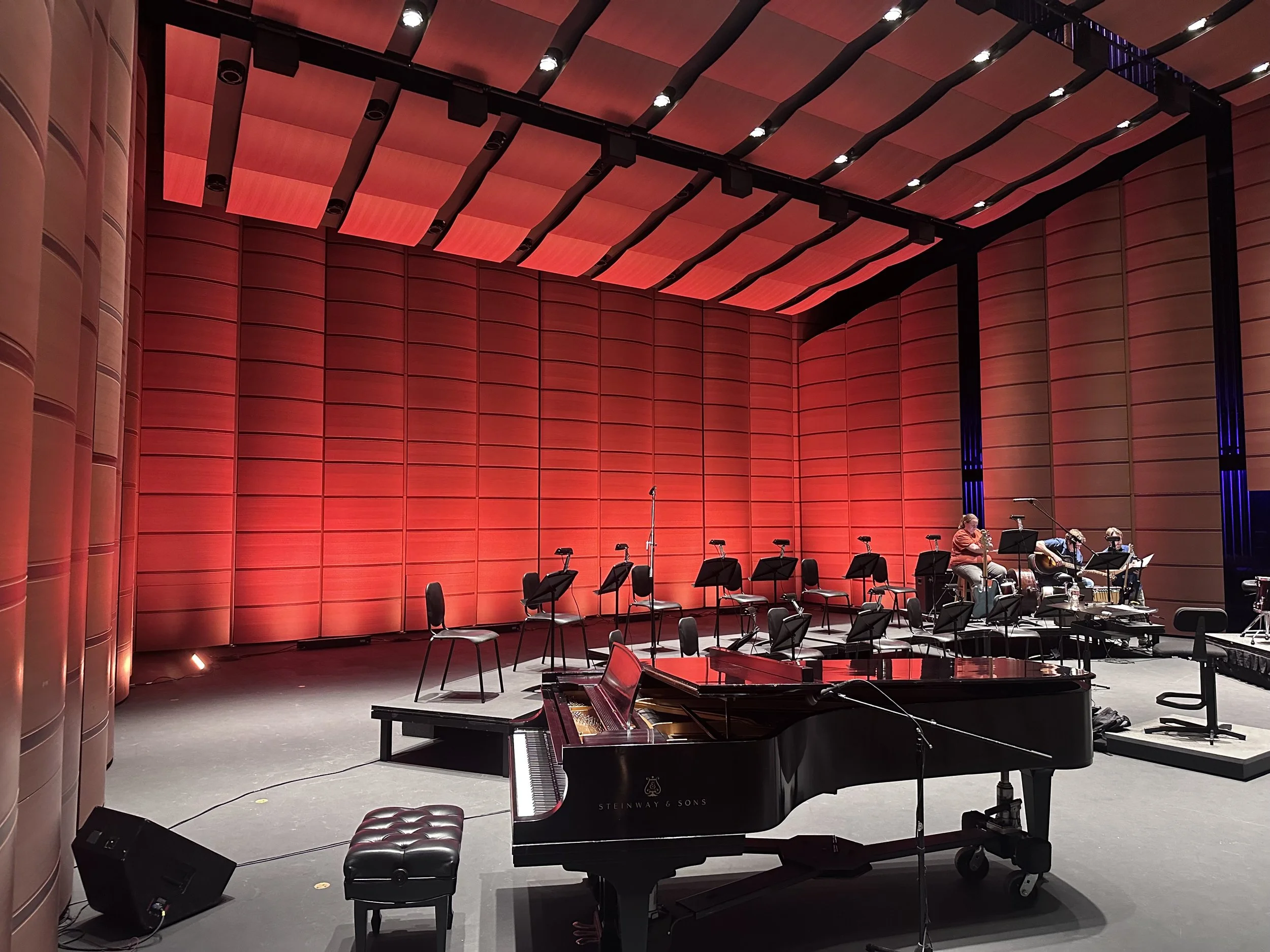 Empty stage of a concert hall with red wall panels, a black grand piano in the foreground, and musicians rehearsing in the background.