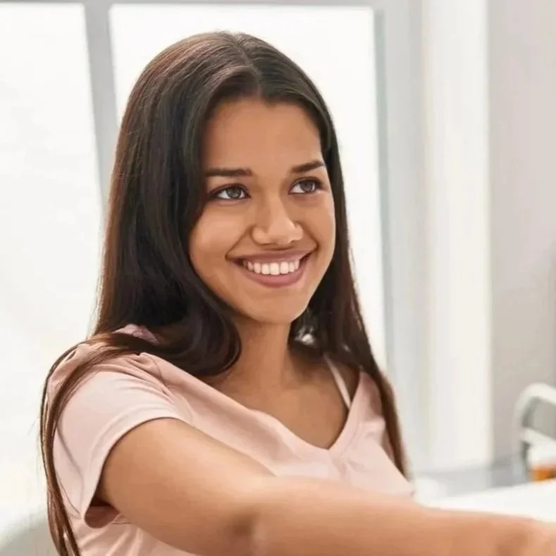 A young woman with long brown hair smiling happily, wearing a light pink shirt in a bright room.