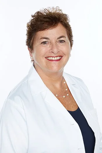 A woman with short curly brown hair wearing a white coat and a navy blue top, smiling against a plain white background.