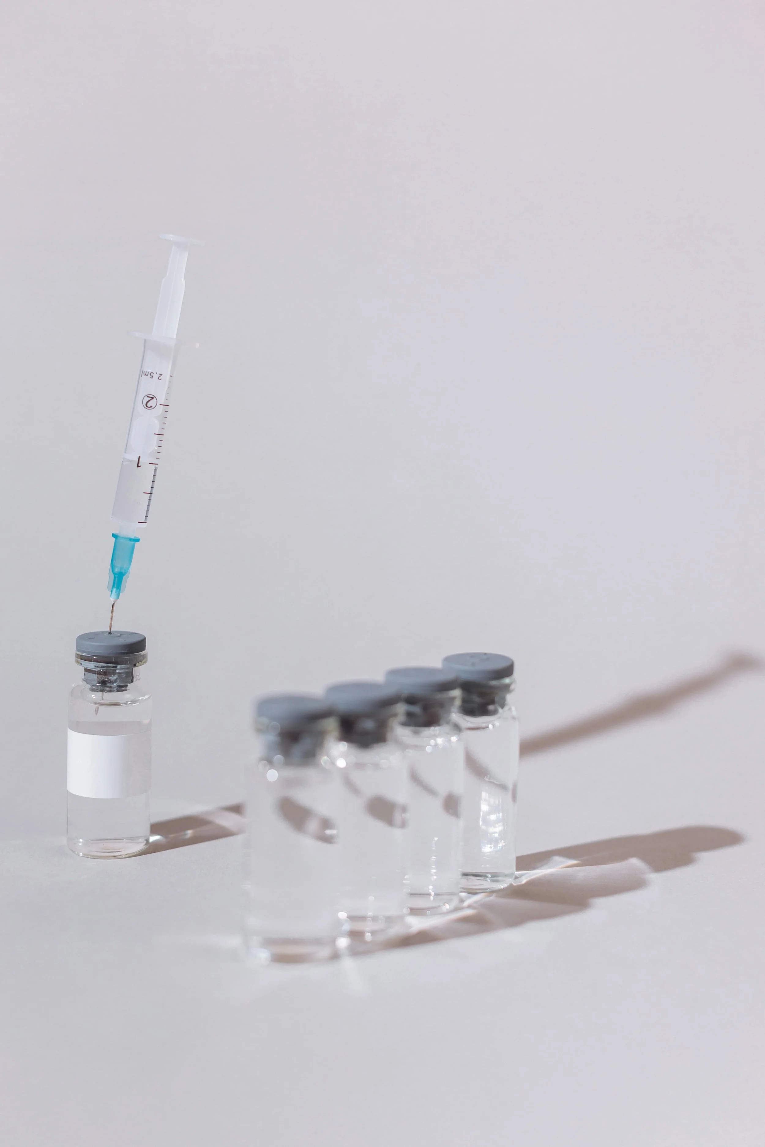 A syringe is drawing a clear liquid from a small vial, with additional vials of clear liquid lined up on a white surface.