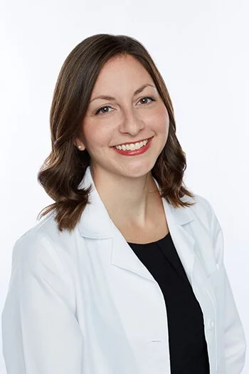 A woman with shoulder-length brown hair, smiling, wearing a white lab coat over a black top against a plain white background.