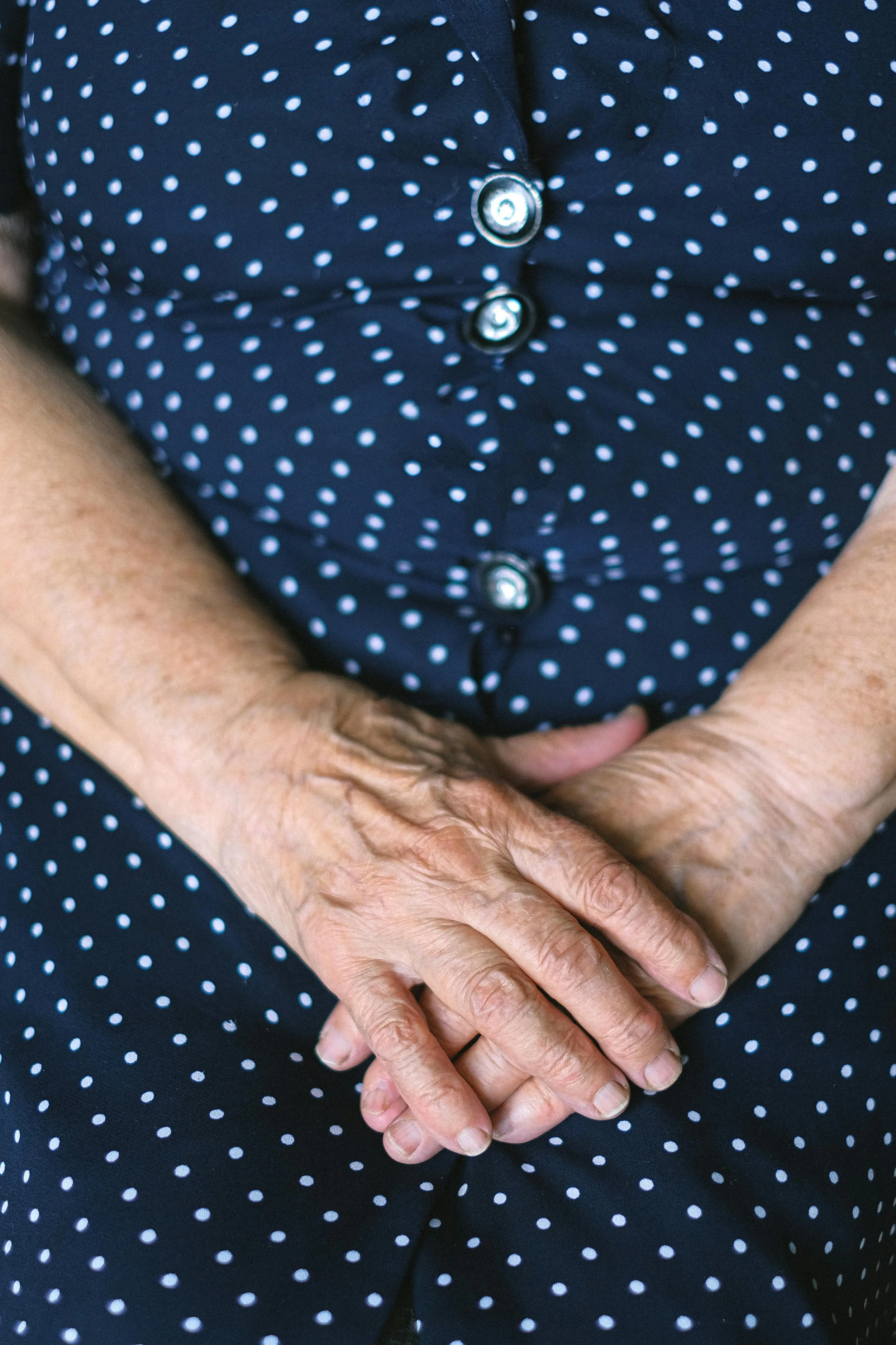 Close-up of elderly person's hands resting on their lap, wearing a navy blue dress with white polka dots and decorative buttons.