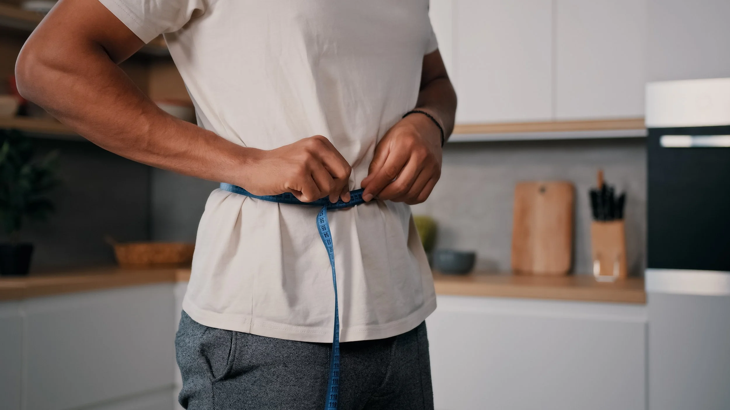 Person measuring their waist with a blue measuring tape in a modern kitchen.