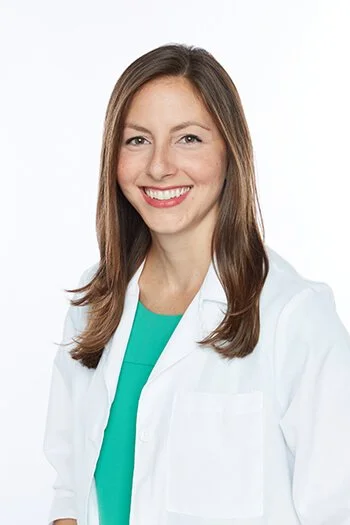 A smiling woman in a white lab coat and teal shirt against a white background.
