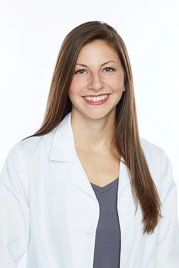 Portrait of a young woman with long brown hair, wearing a white lab coat and gray top, smiling against a plain white background.