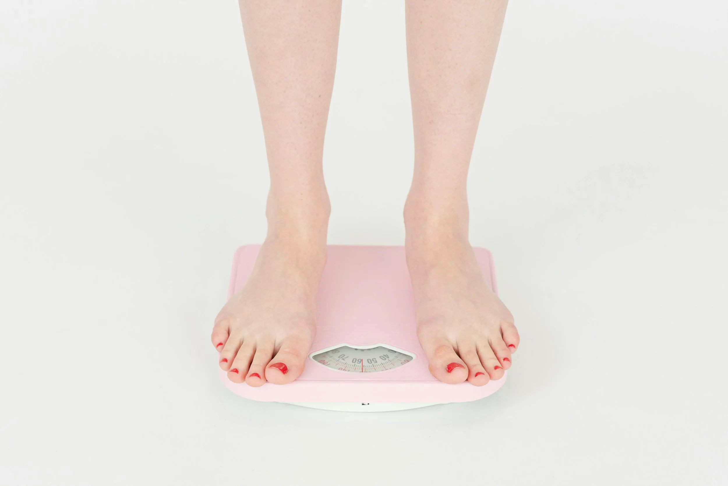 Close-up of a woman standing barefoot on a pink mechanical bathroom scale, measuring her weight, with a white background.