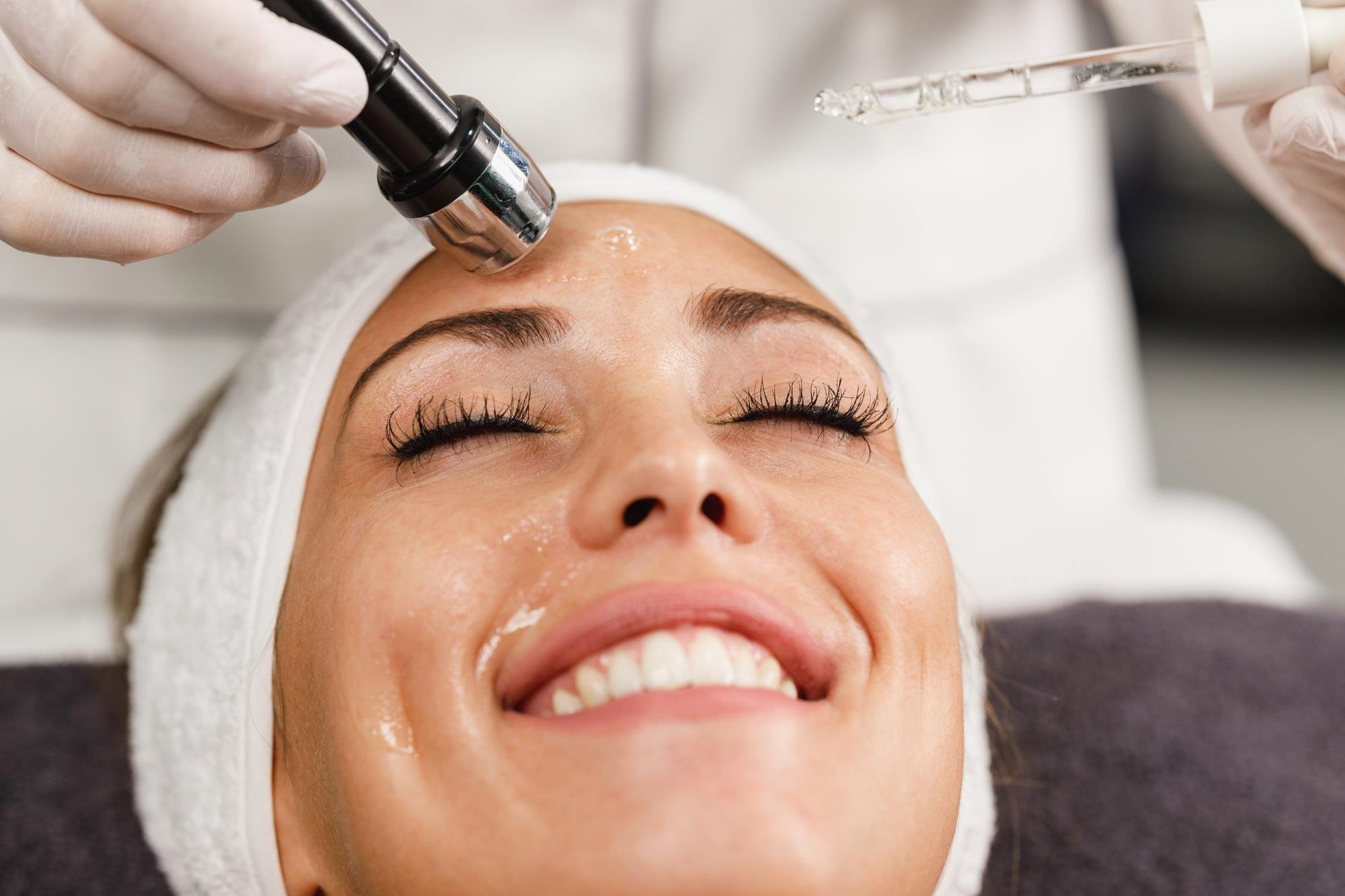 Beauty professional performing facial treatment on woman with closed eyes, smiling, lying on treatment bed with head wrapped in white towel.