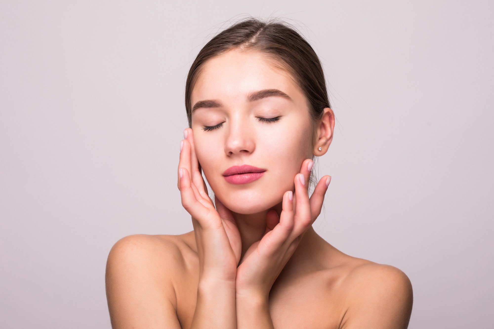 A young woman with closed eyes and flawless skin gently touching her face with both hands.