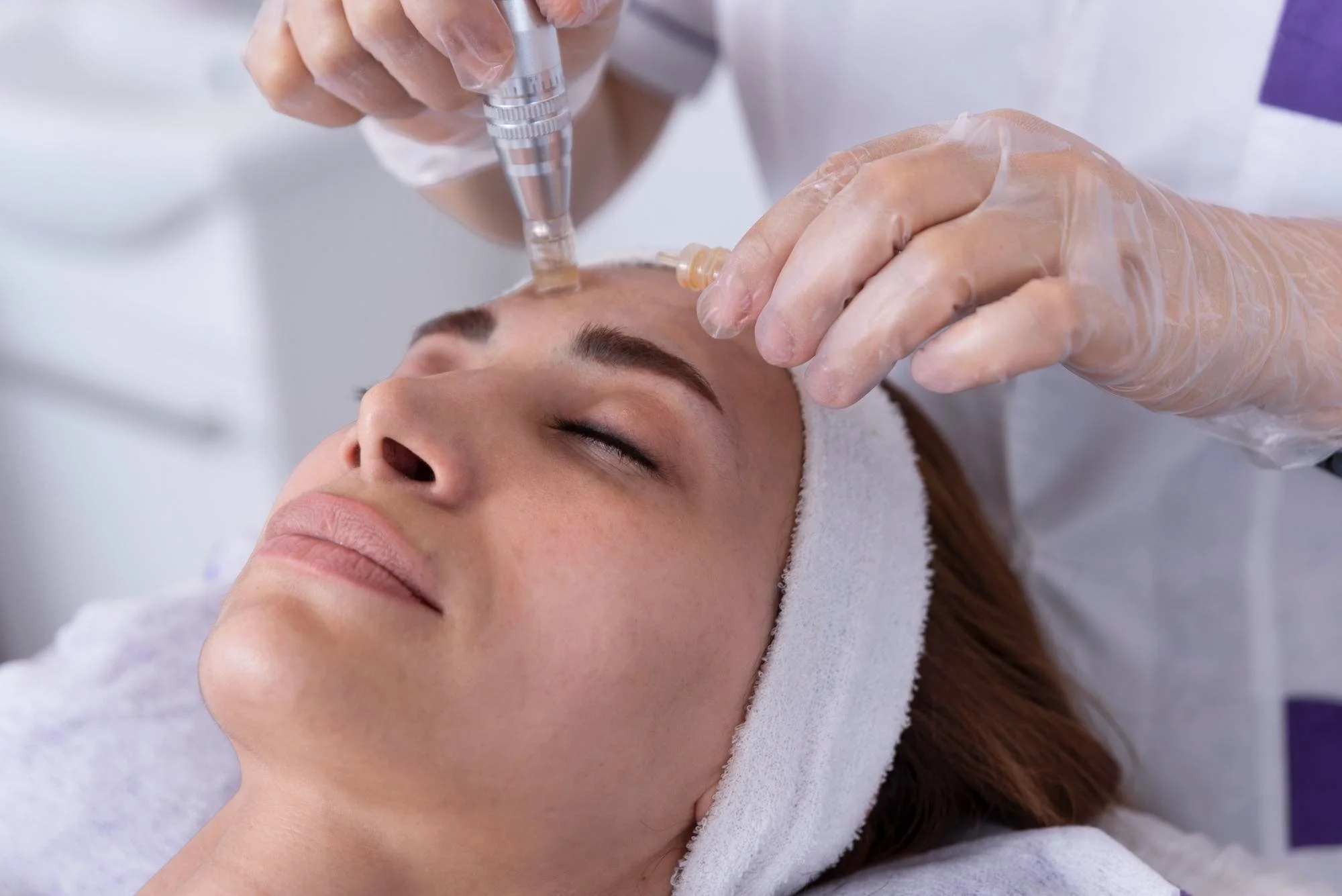 A woman receives a facial treatment with a procedure that appears to involve a syringe or applicator, while lying down with her eyes closed in a spa or skincare clinic.
