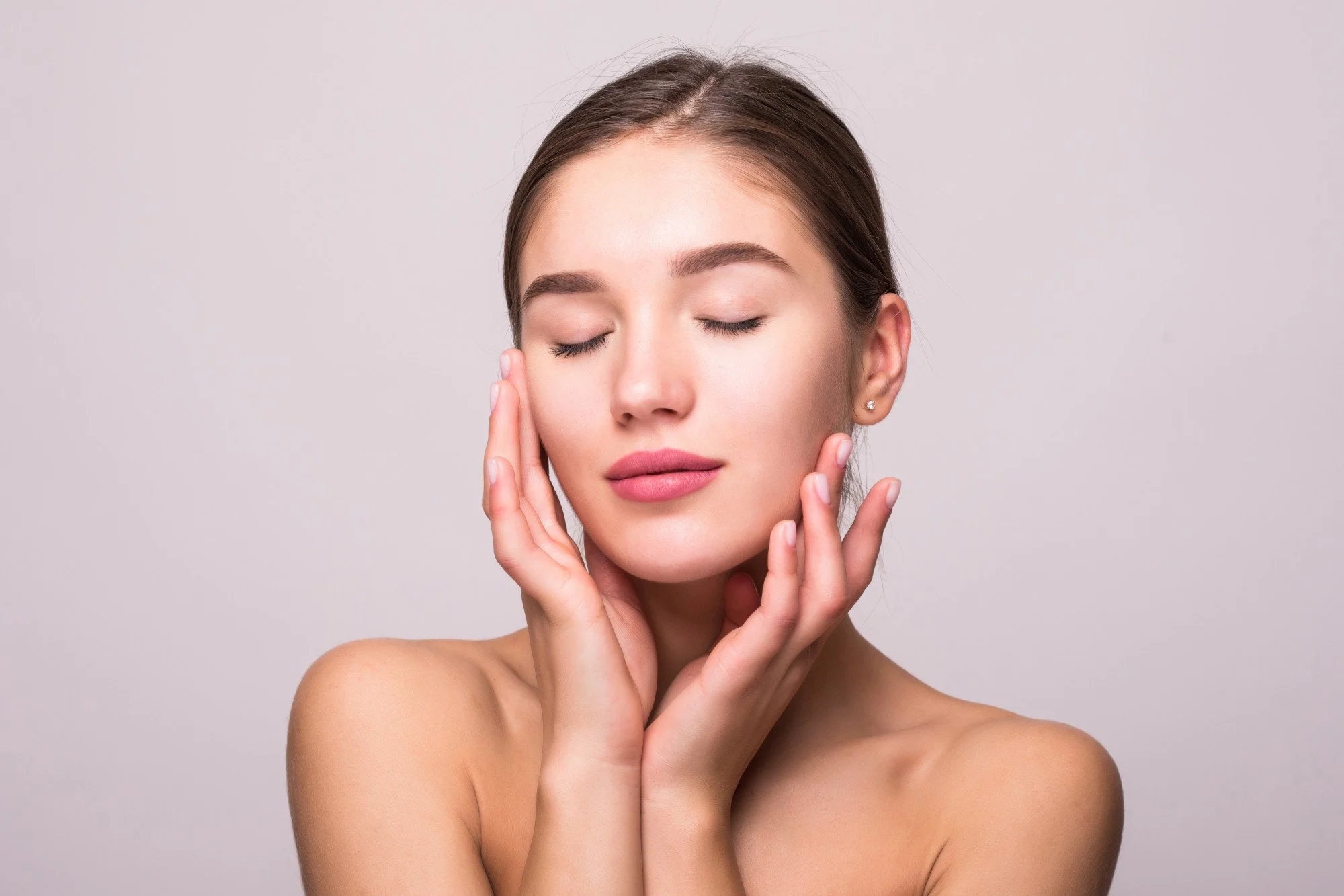 A young woman with smooth skin and closed eyes touching her face with her hands, standing against a plain light gray background.
