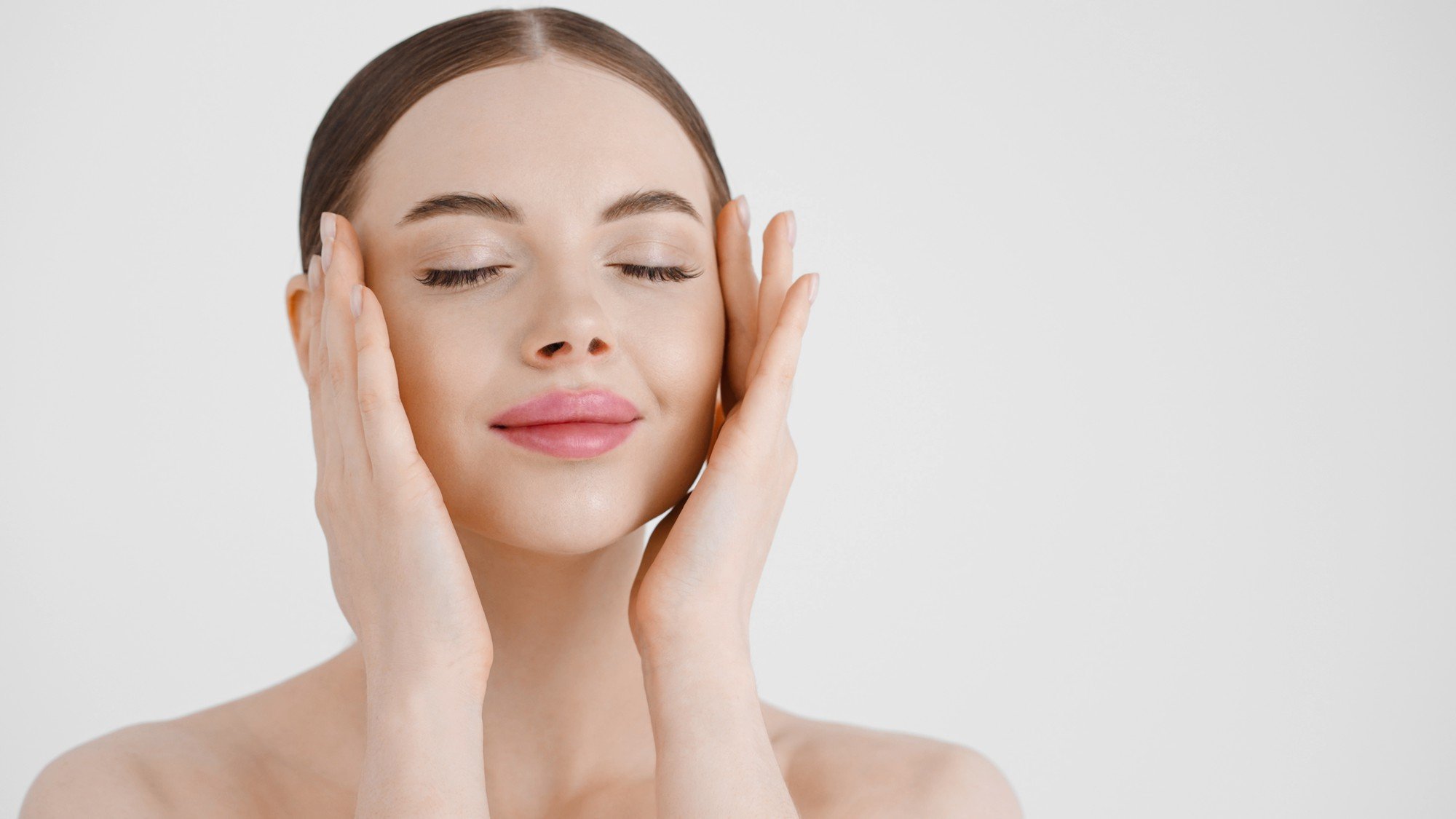 A young woman with closed eyes and a serene expression gently holds her face with both hands.