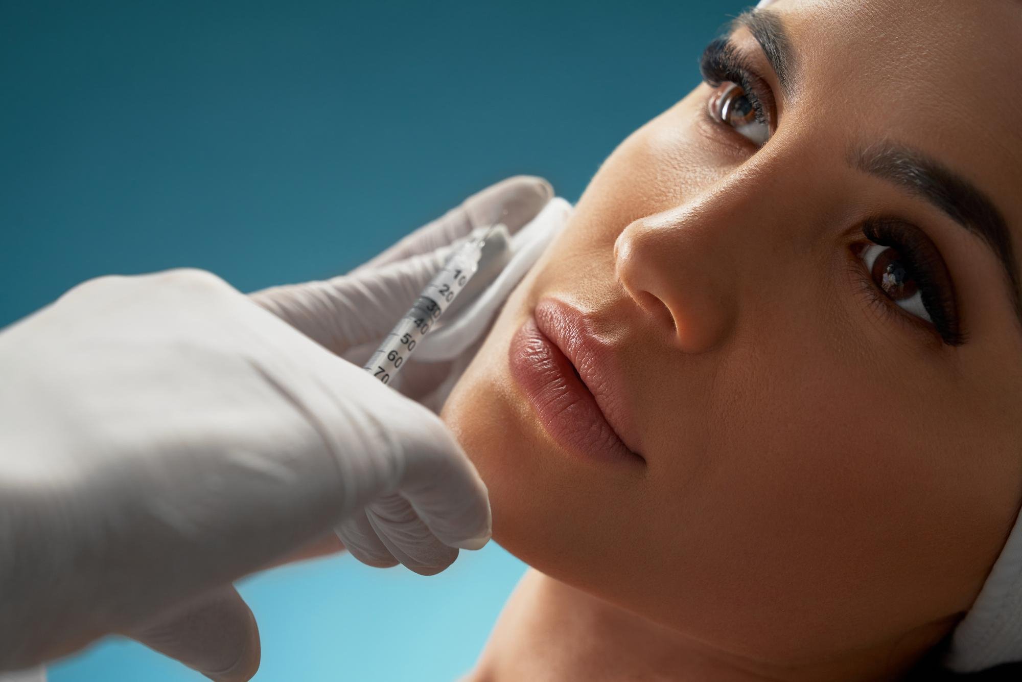 A healthcare professional wearing white gloves is administering an injection into a woman's upper arm, who is lying down with her face close to the camera, against a blue background.