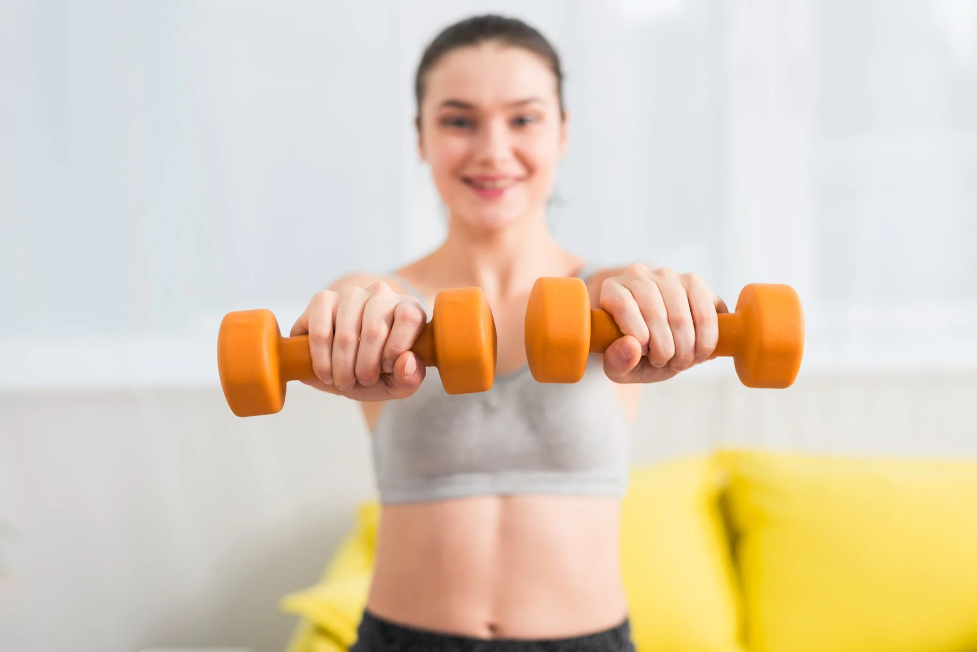 A woman smiling and holding an orange dumbbell towards the camera in a bright, minimalistic workout space.