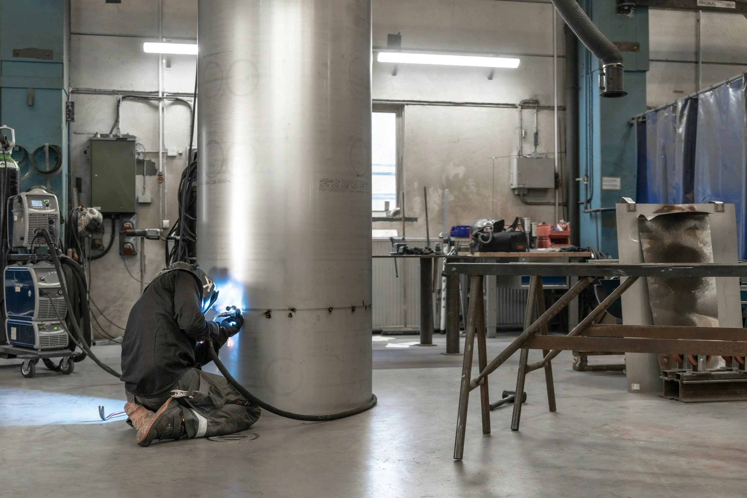A worker is welding a large metal cylinder inside a workshop or industrial space, with welding equipment nearby and various tools and machinery around.
