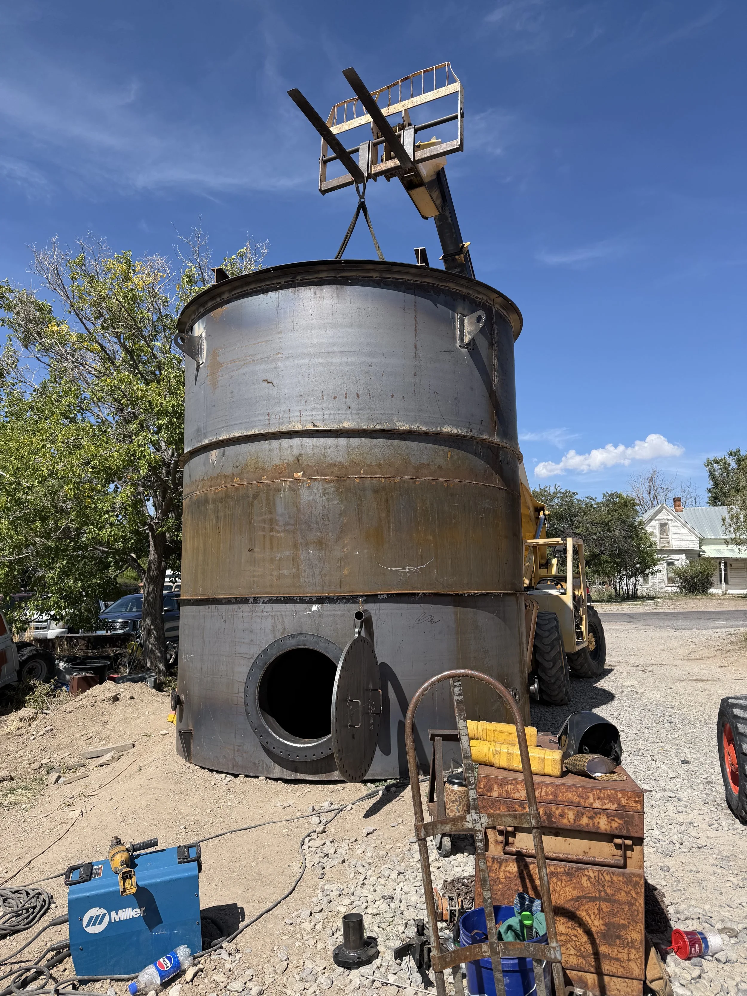 A massive custom-fabricated carbon steel storage tank with a bottom manway access, being positioned by heavy equipment outside the Peak Welding shop.