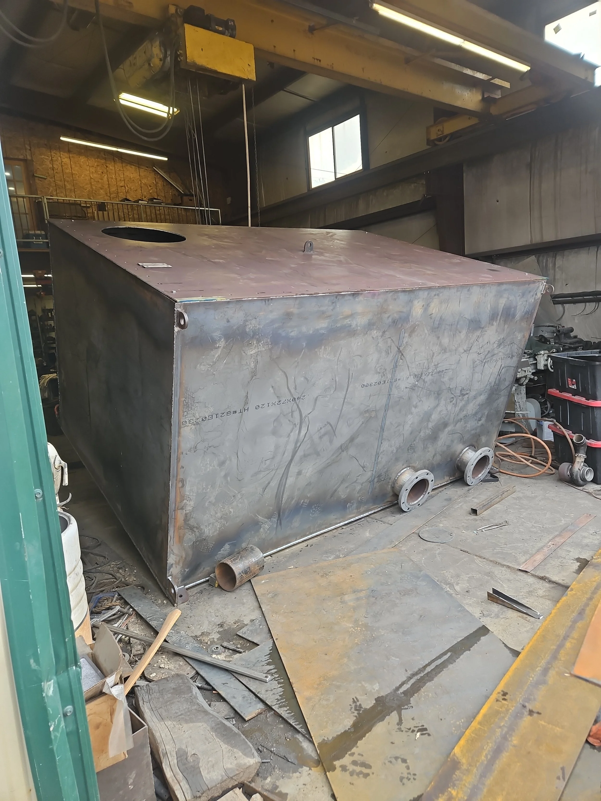 A large fabricated carbon steel industrial tank with flanged connections resting on the shop floor under an overhead crane inside Peak Welding's Manti, Utah facility.