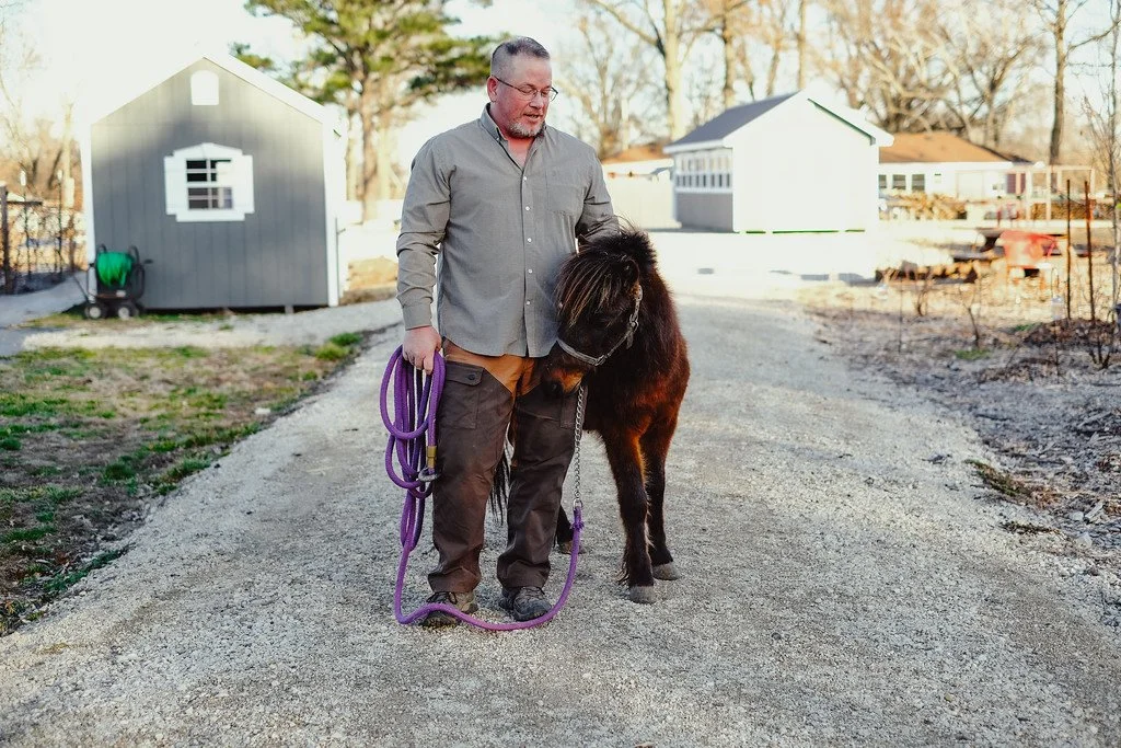 A man holding a purple leash stands on a gravel path with a small dog, in an outdoor setting with small buildings and trees in the background.