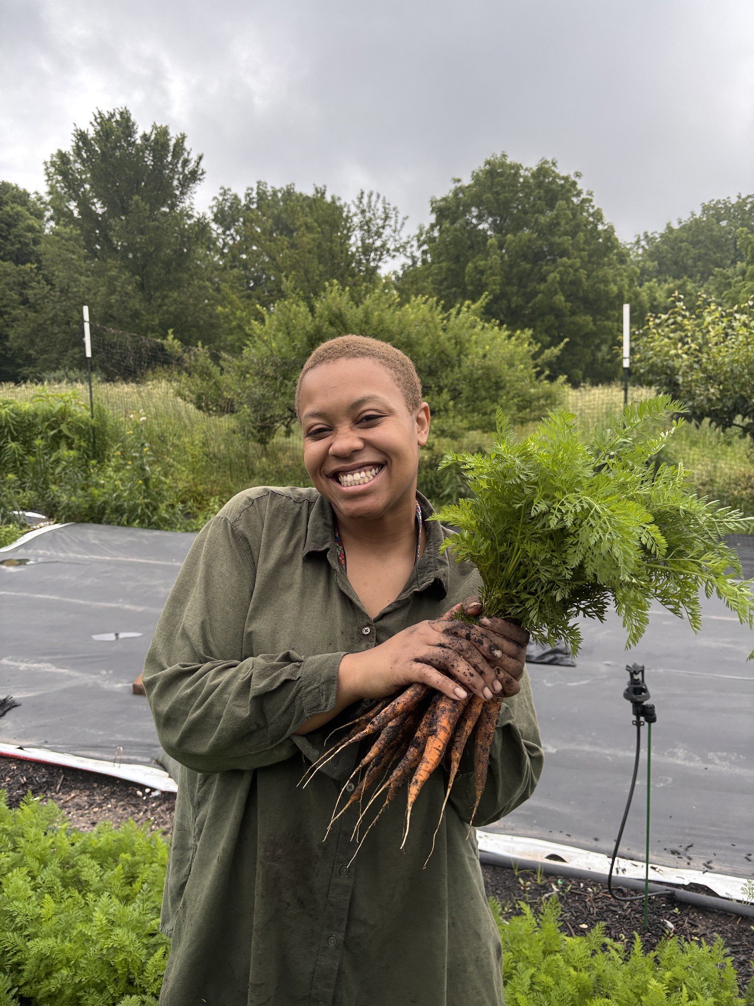 A woman smiling and holding freshly harvested carrots in a garden with greenery and trees in the background.