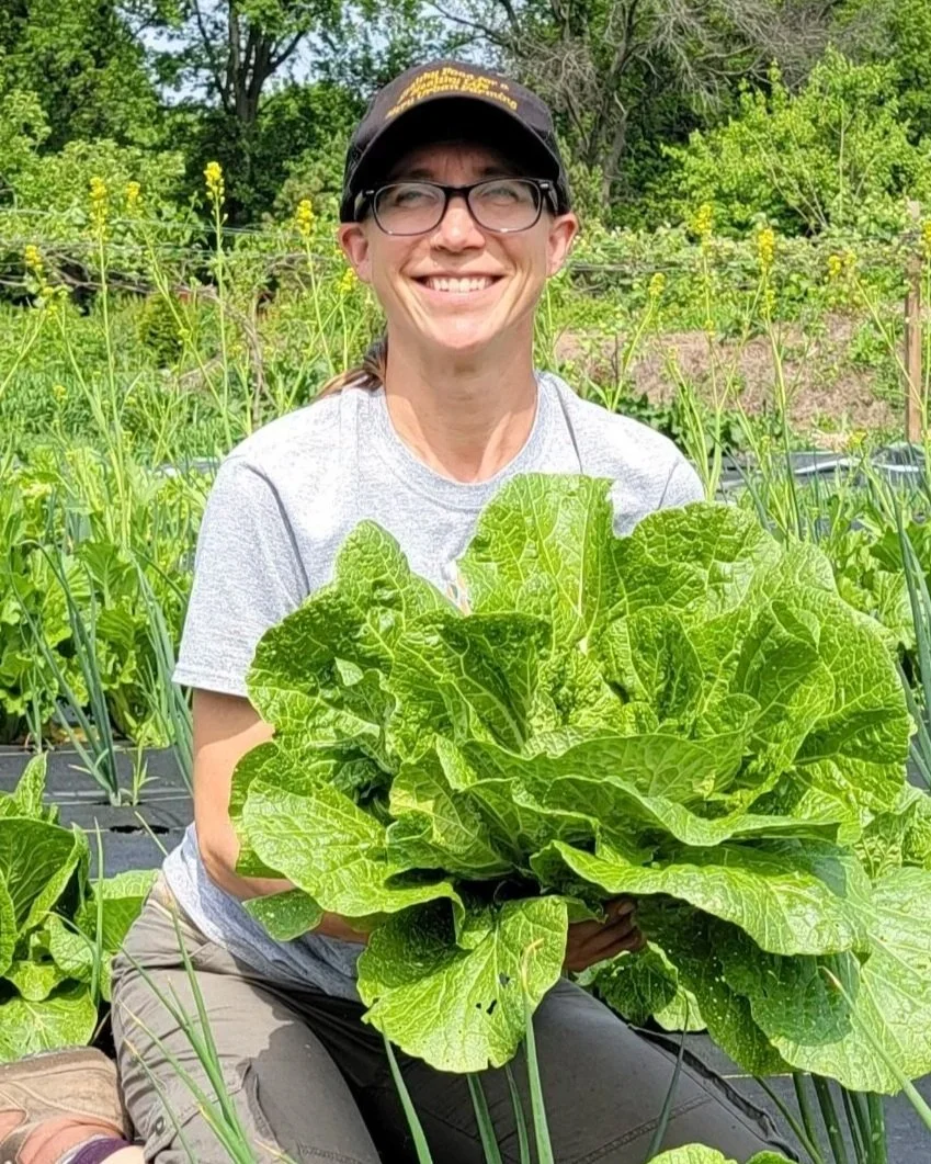A smiling woman wearing glasses, a gray t-shirt, and a black cap, holding a large leafy green plant in a garden with flowering plants and trees in the background.
