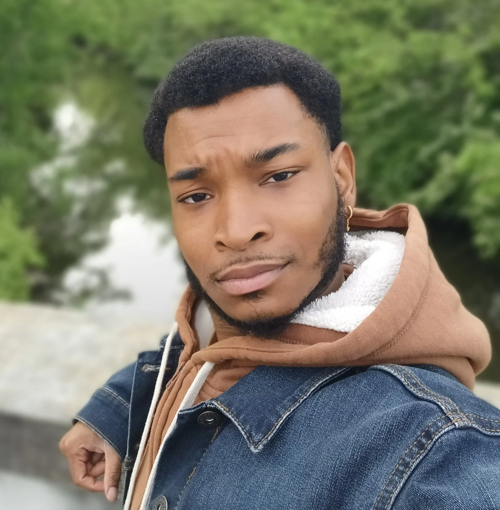 A young man with dark hair and facial hair, wearing a blue denim jacket over a brown hoodie and white inner lining, taking a selfie outdoors with greenery and a body of water in the background.