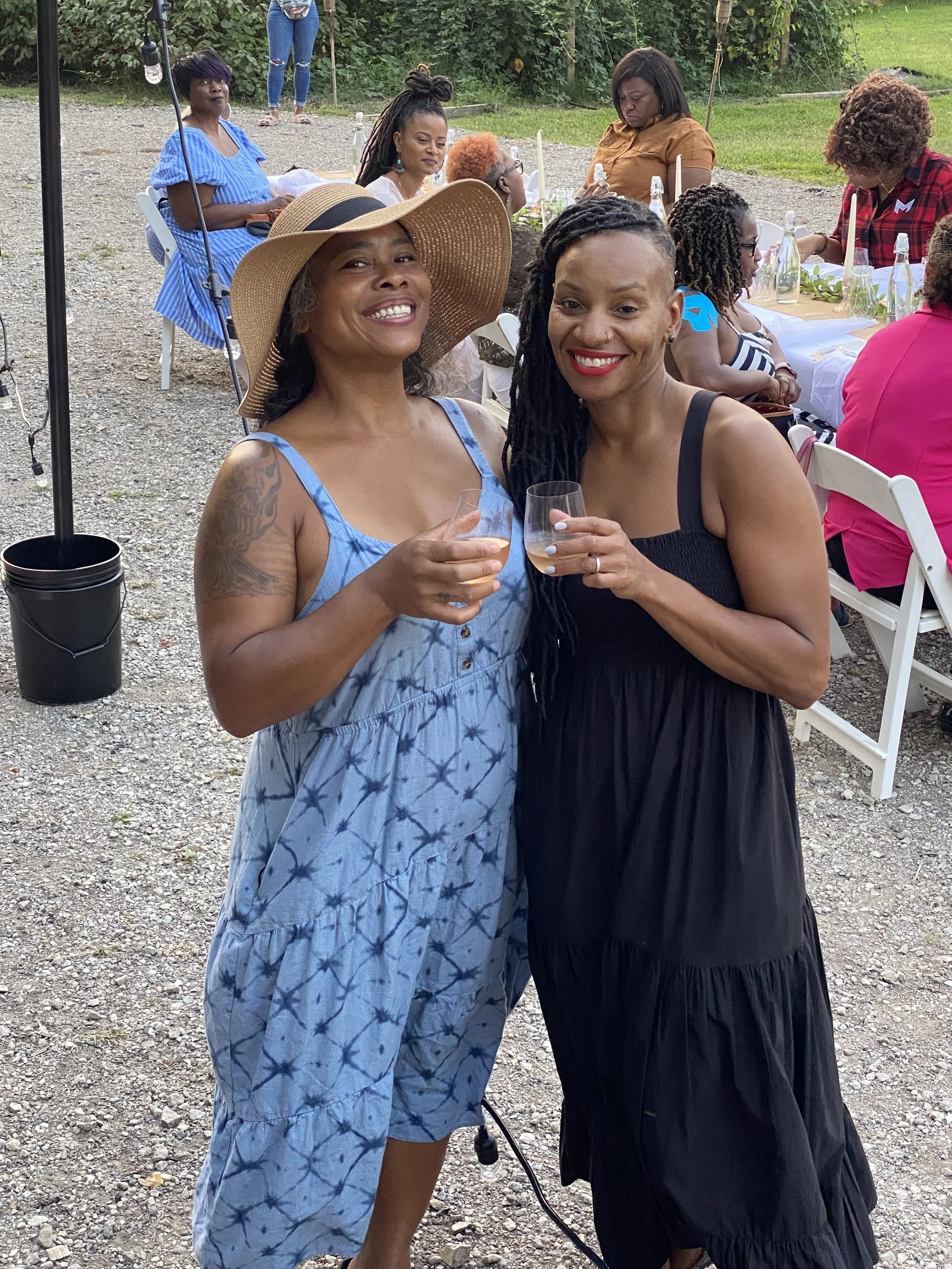 Two women holding glasses of drink, smiling outdoors at a gathering with other women seated at a table in the background.