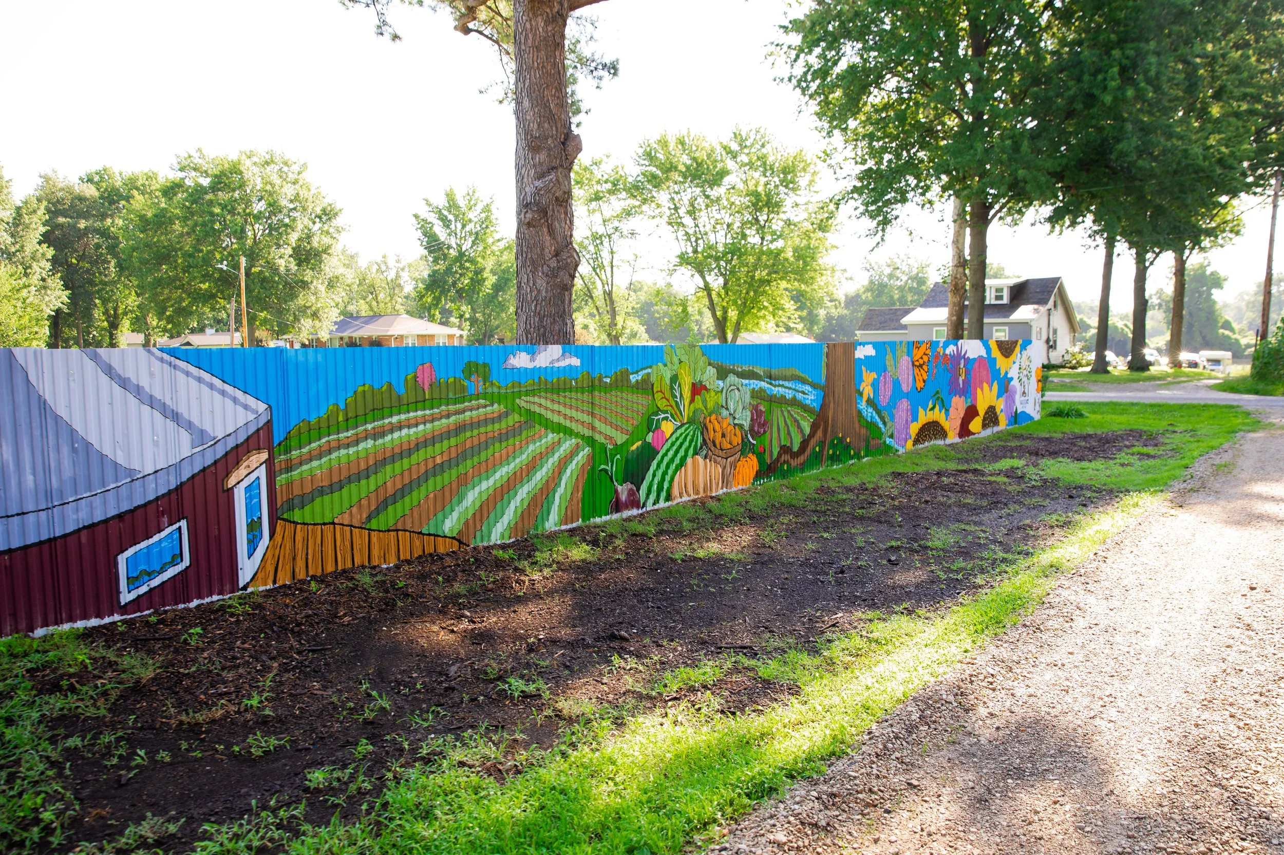Colorful mural painted on a curved metal fence depicting a farm landscape with fields, trees, and flowers, located next to a dirt pathway and green grass, with tall trees and houses in the background.