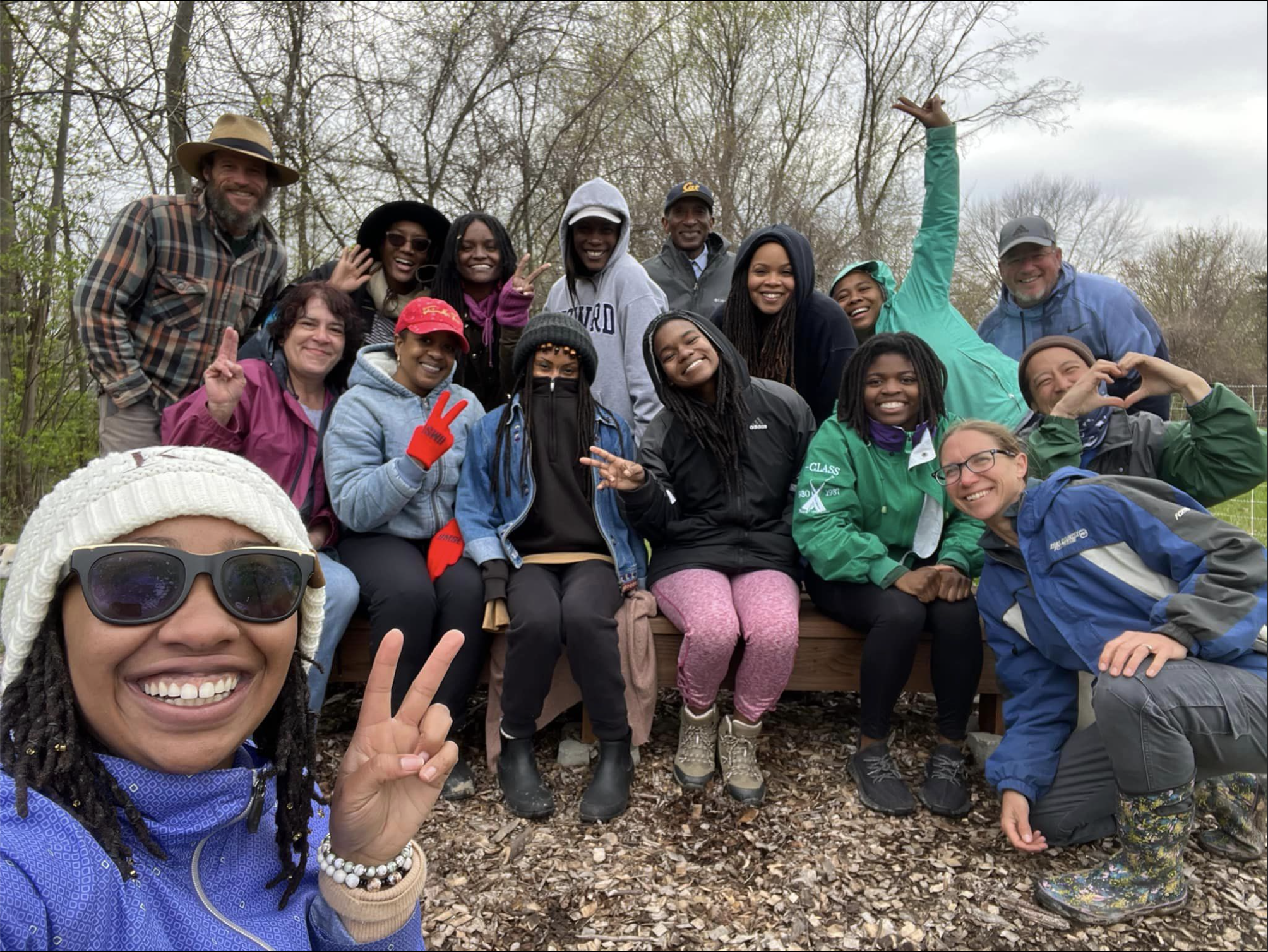 A group of diverse friends outdoors, smiling and posing for a photo with trees and cloudy sky in the background.