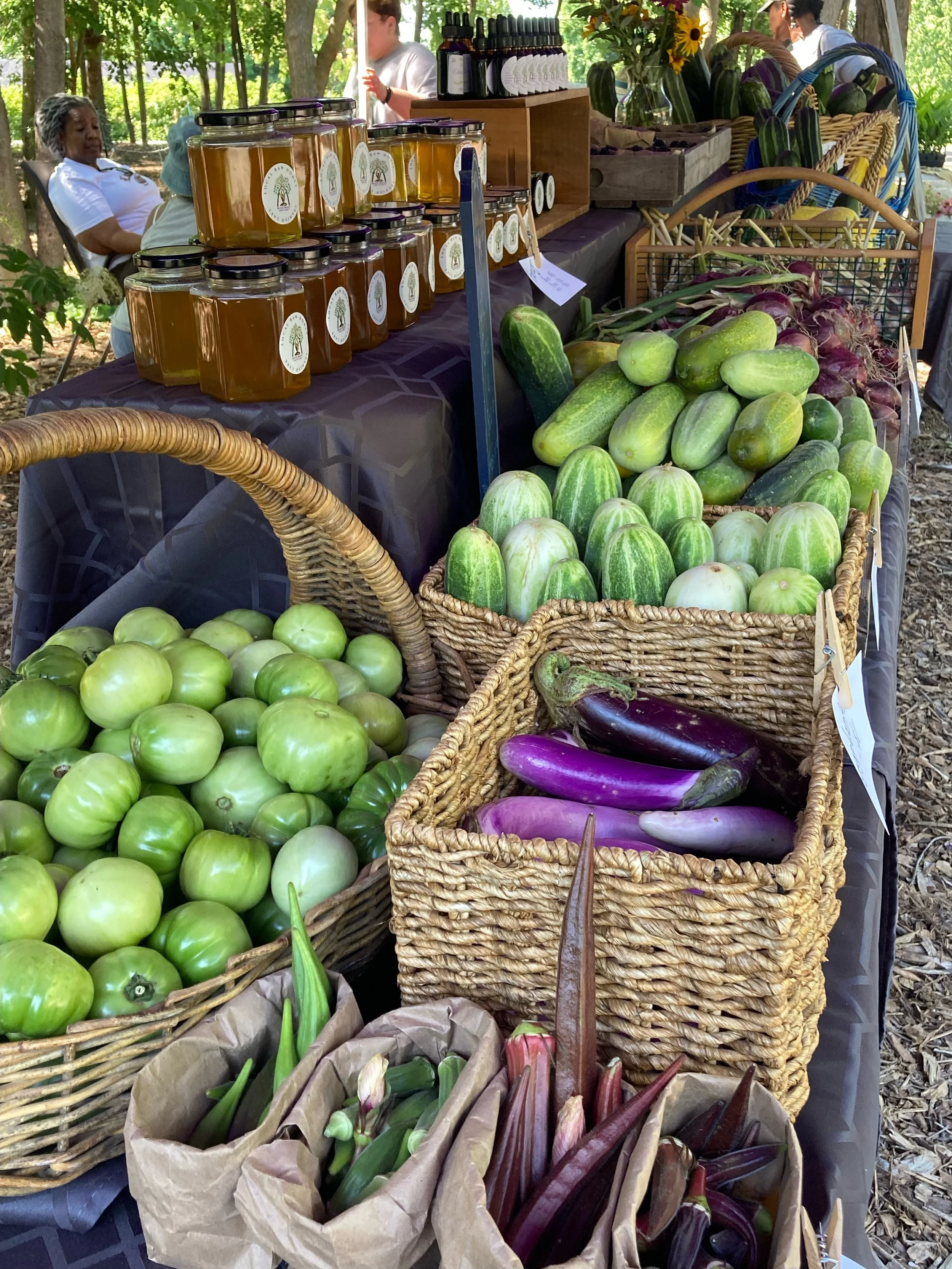 Display of fresh vegetables including tomatoes, eggplants, zucchini, and onions at an outdoor market stand with jars of honey and bottles.