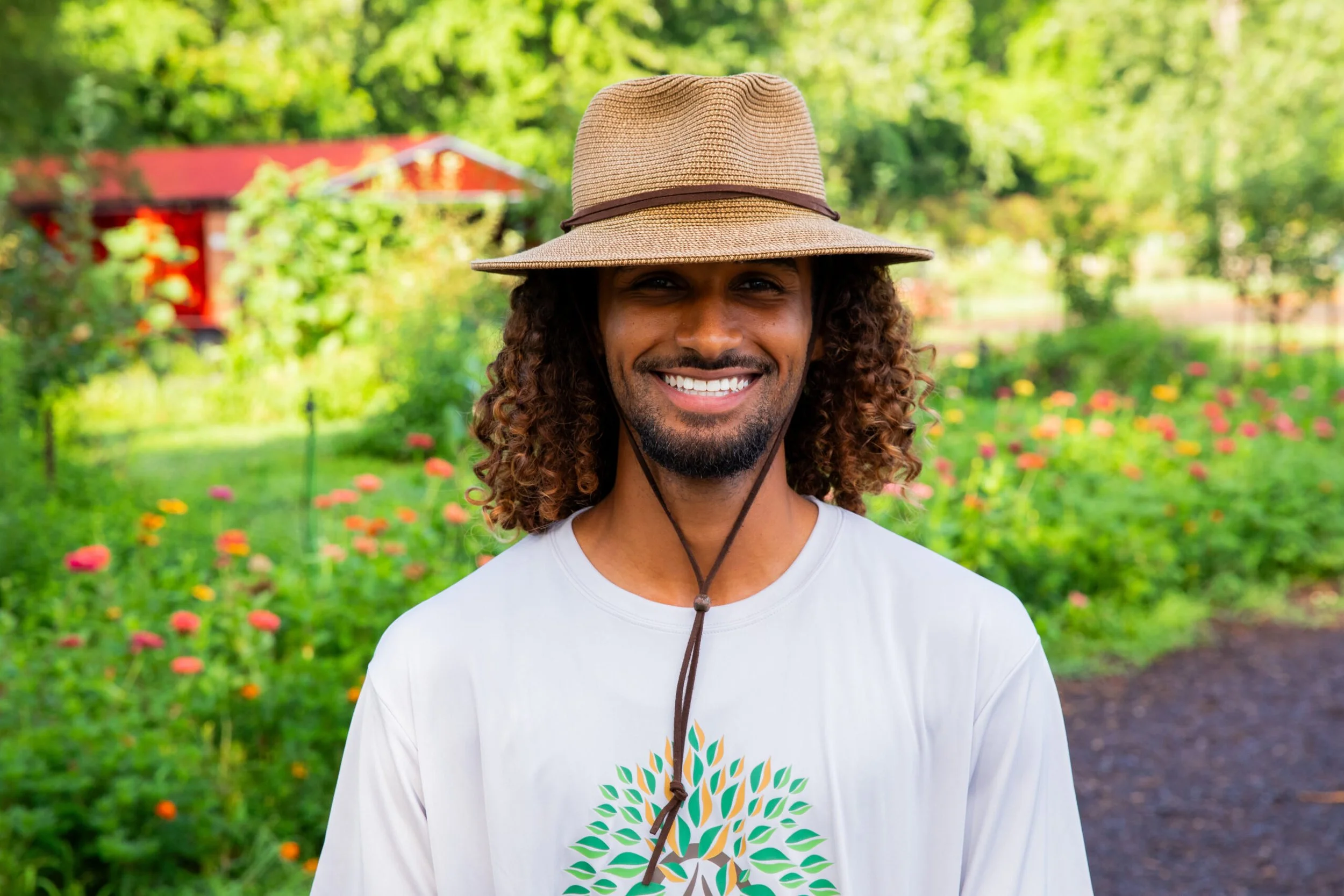 Smiling man with curly hair wearing a wide-brimmed hat standing outdoors in a garden with flowers and greenery.