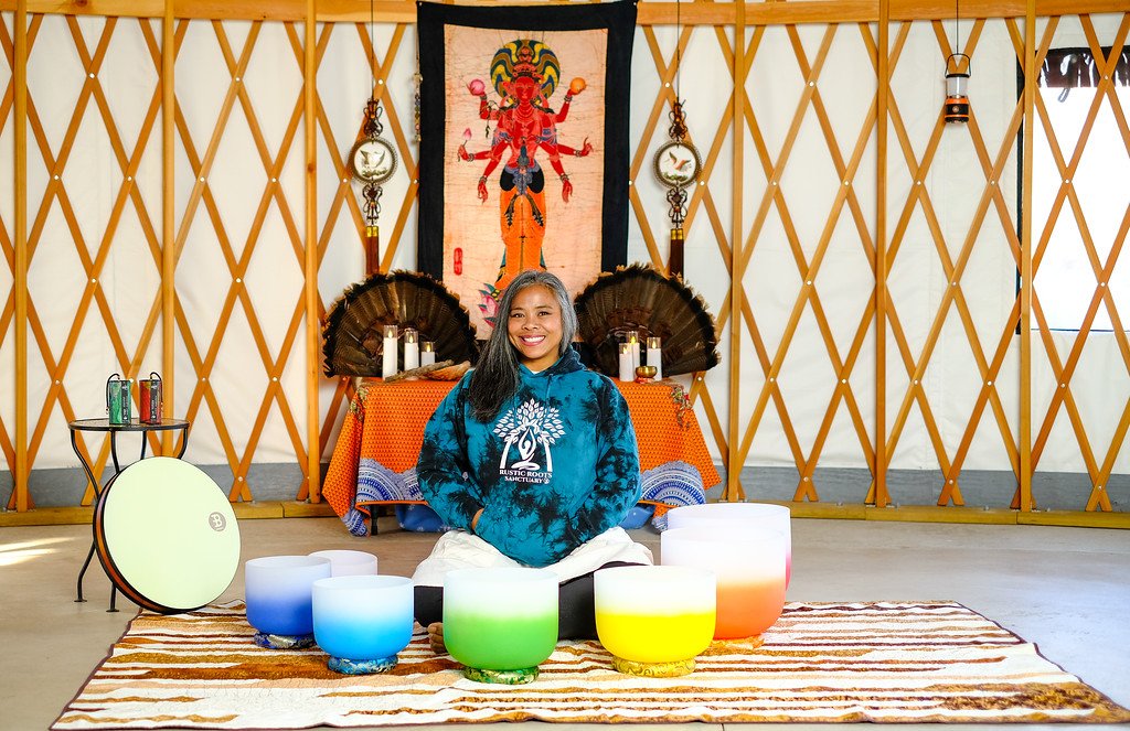 A woman sitting cross-legged on a rug surrounded by colorful singing bowls in a yurt with Native American decor and spiritual symbols.