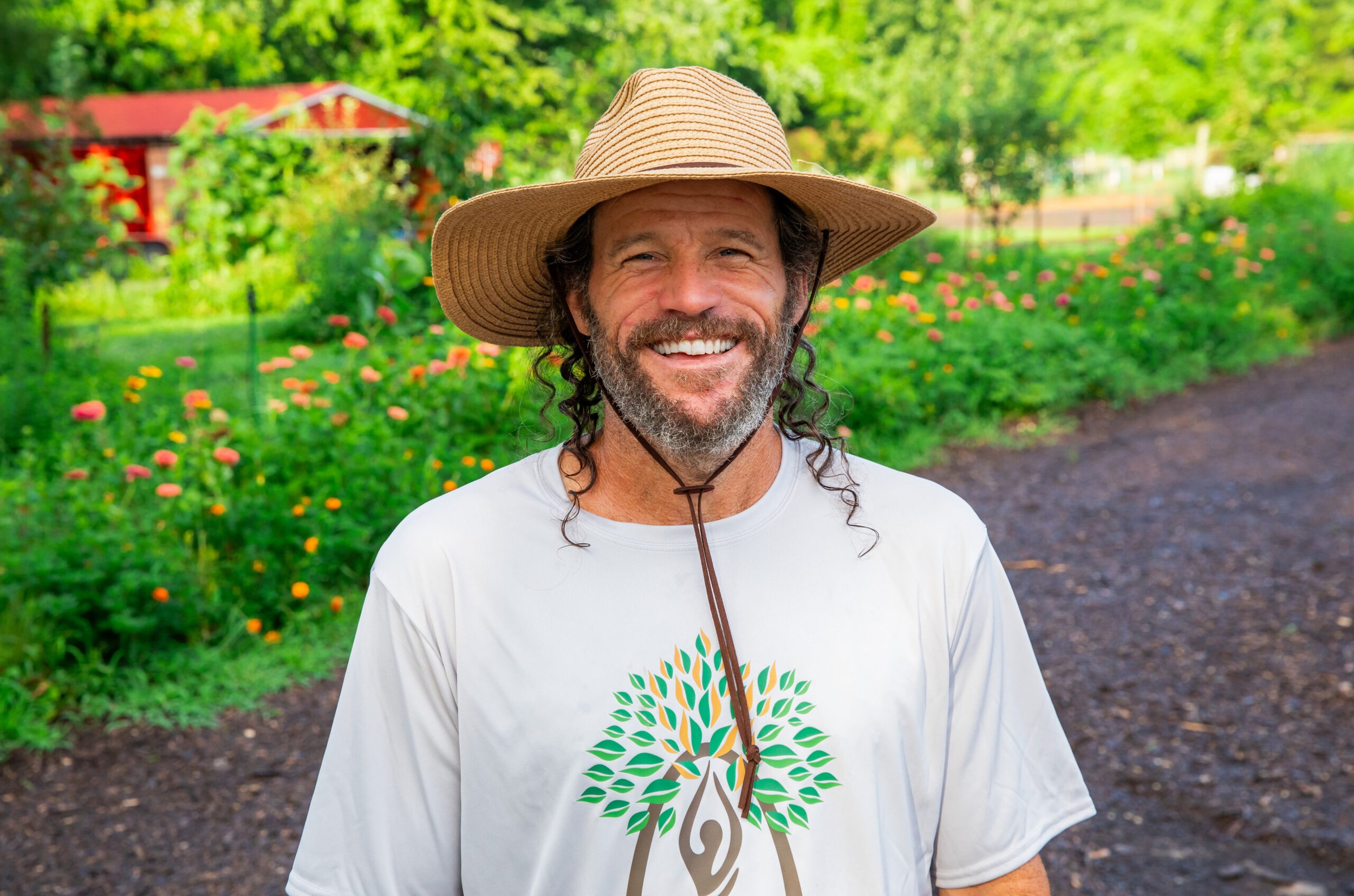 Smiling man wearing a wide-brimmed hat and a white t-shirt with a tree logo stands outdoors in a garden with flowers and green trees.