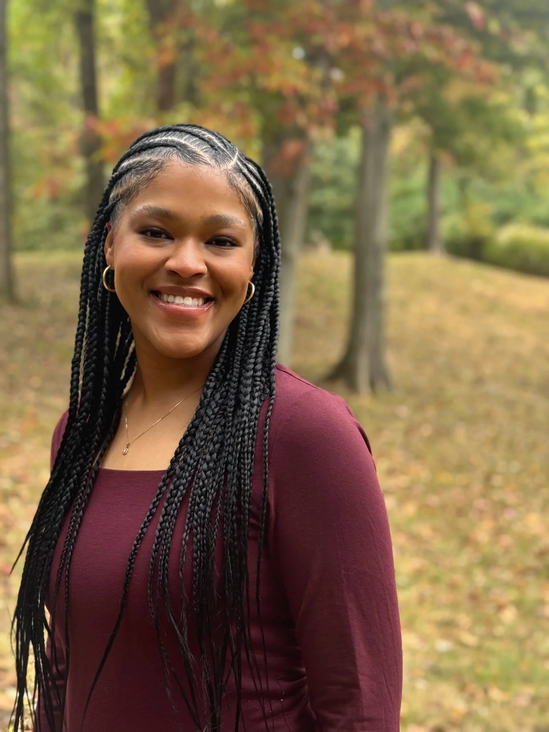 A young woman with braided hair, wearing a maroon long-sleeve top, smiling outdoors with a background of trees with autumn foliage.