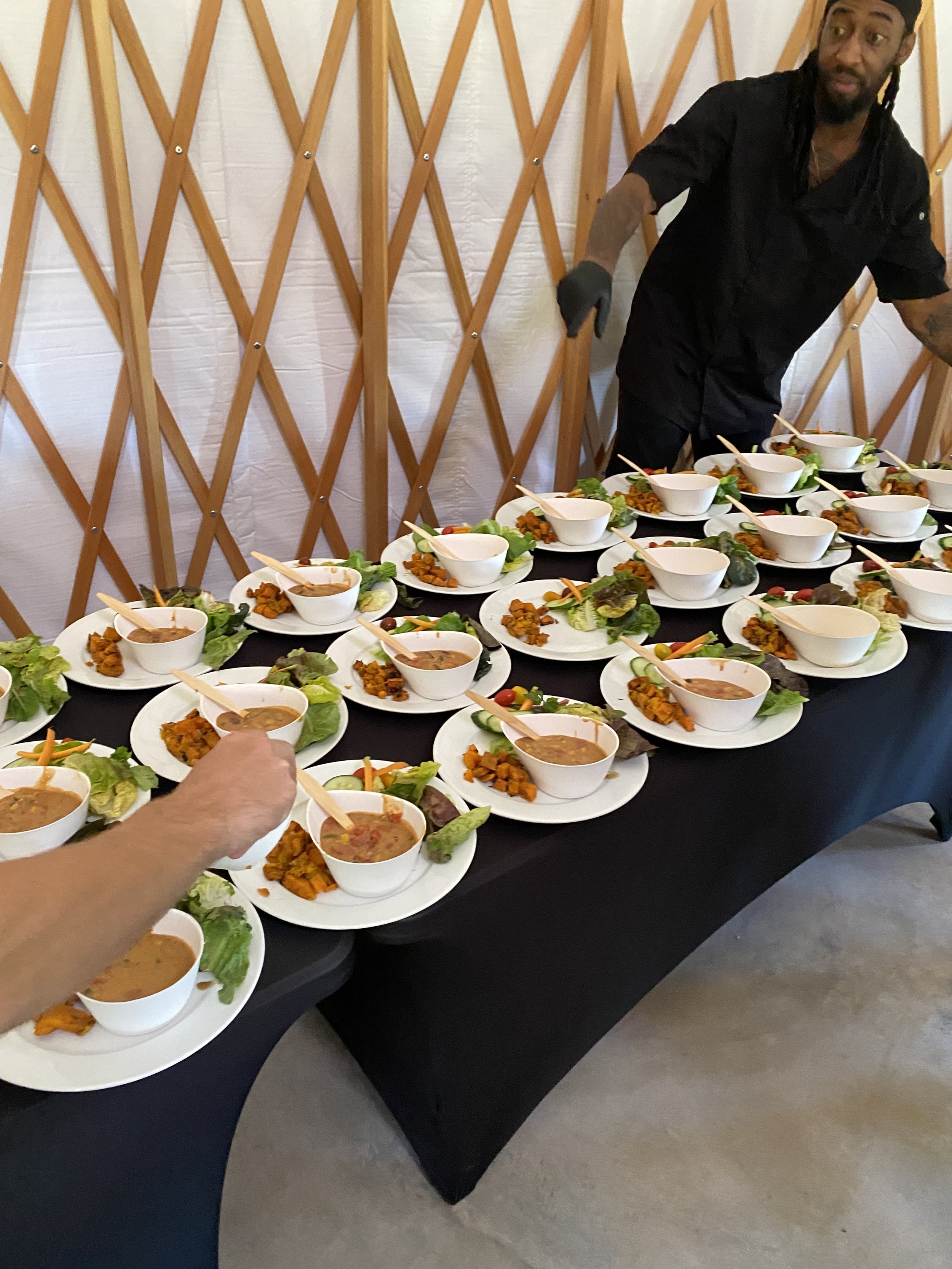 A man wearing a black shirt and black glove standing behind a table with multiple white bowls of food, garnished with vegetables and lettuce leaves, in a setting with a wooden lattice wall in the background.