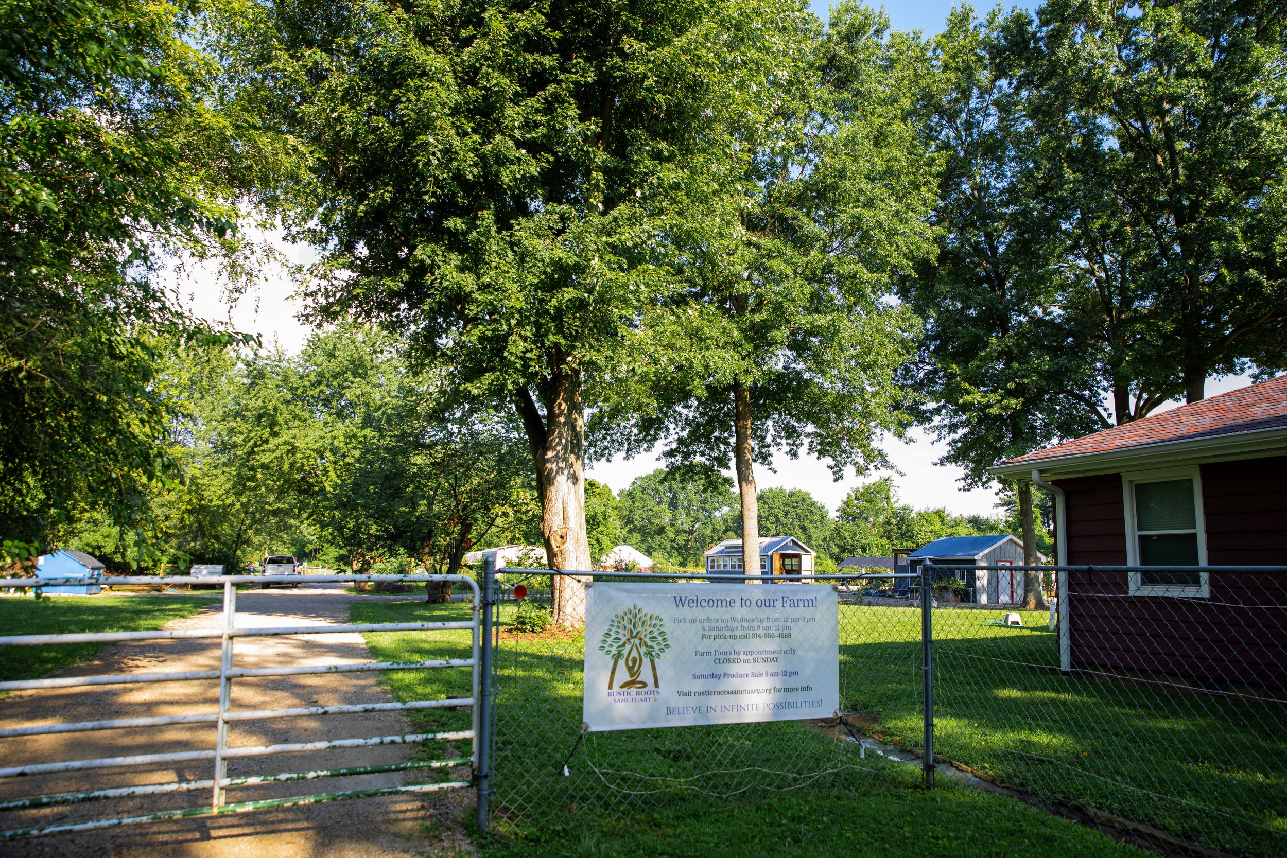 Entrance gate to a farm with a sign welcoming visitors, surrounded by green trees and a grassy area, with farm buildings in the background.