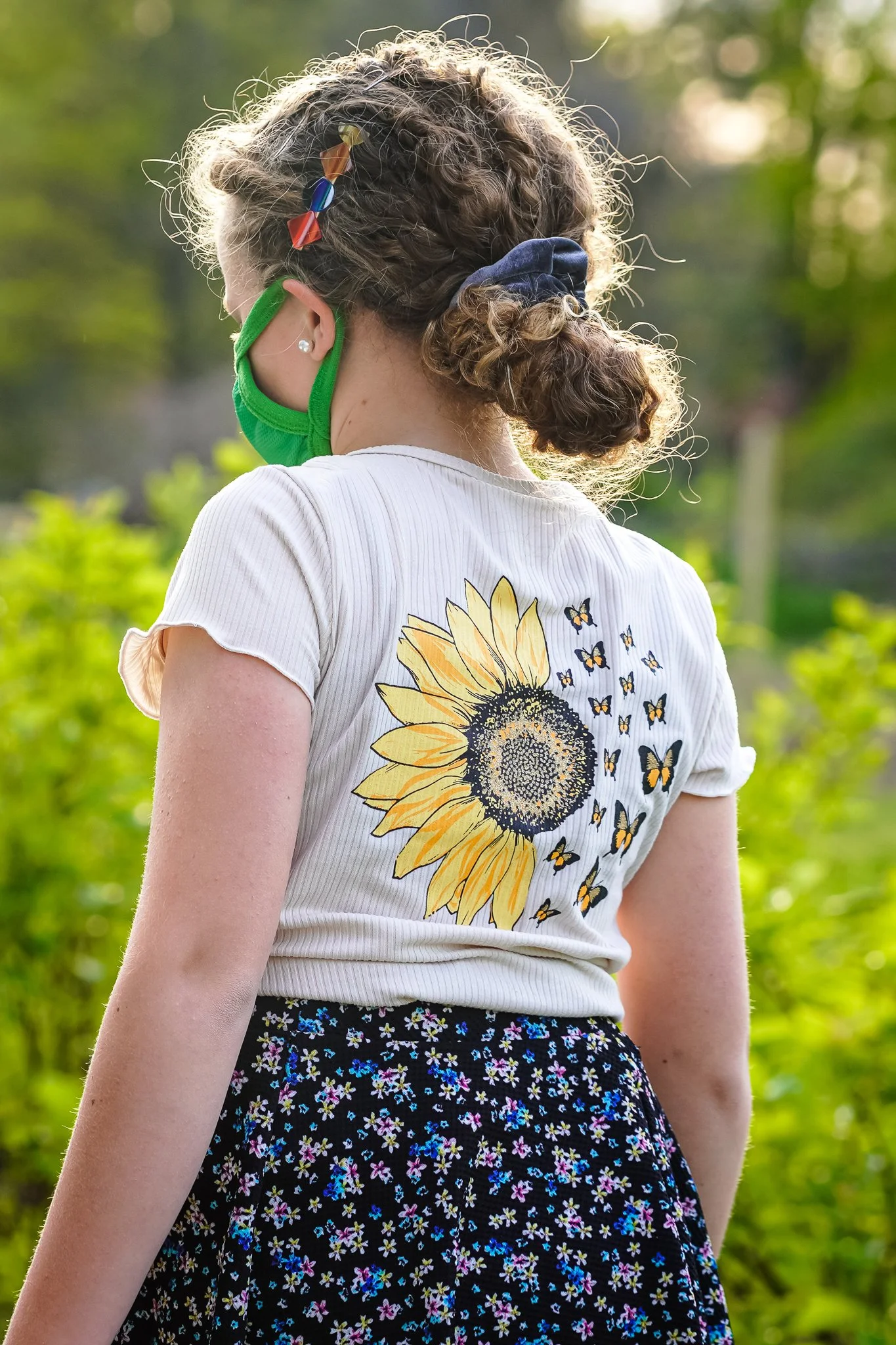 A girl with curly hair tied in a bun wearing a green face mask, a white T-shirt with a large sunflower and butterflies printed on the back, and a black floral skirt, standing outdoors with greenery in the background.