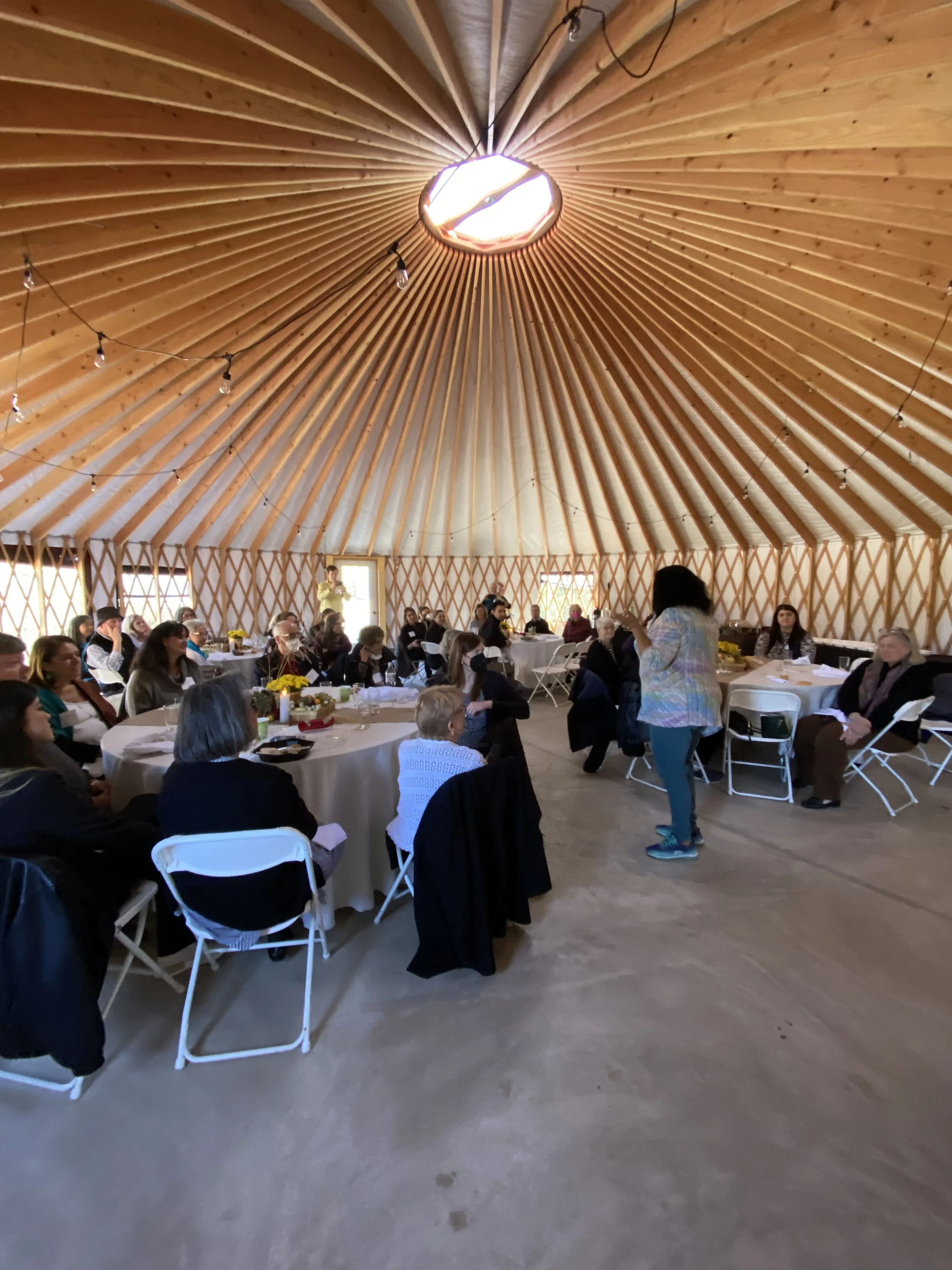 People gathered inside a large wooden round building with a conical ceiling, seated at round tables with white tablecloths, listening to a woman speaking. The ceiling has string lights and a central opening revealing the sky.
