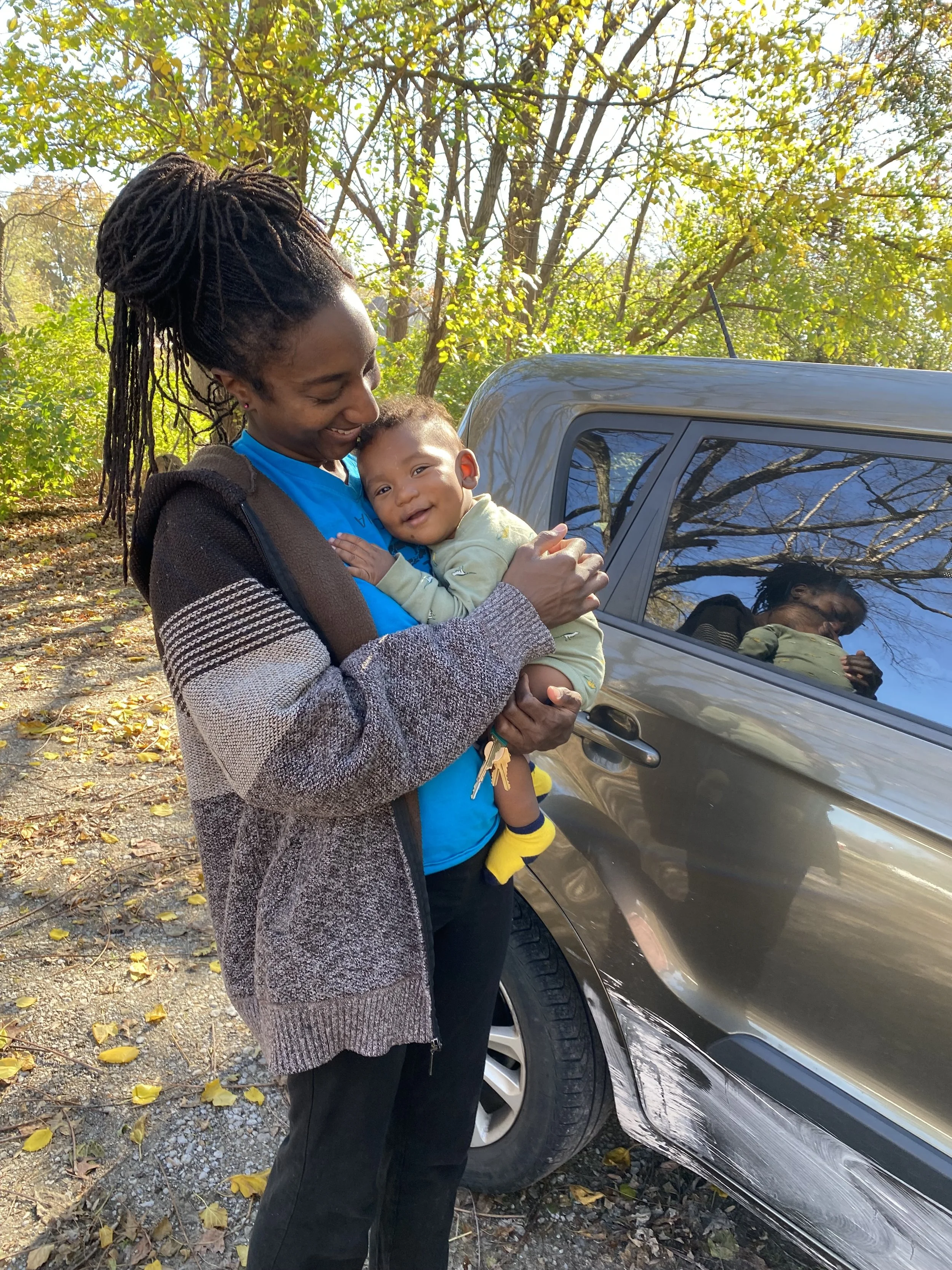 Woman holding smiling baby outdoors near a parked vehicle with trees and fallen leaves in the background.