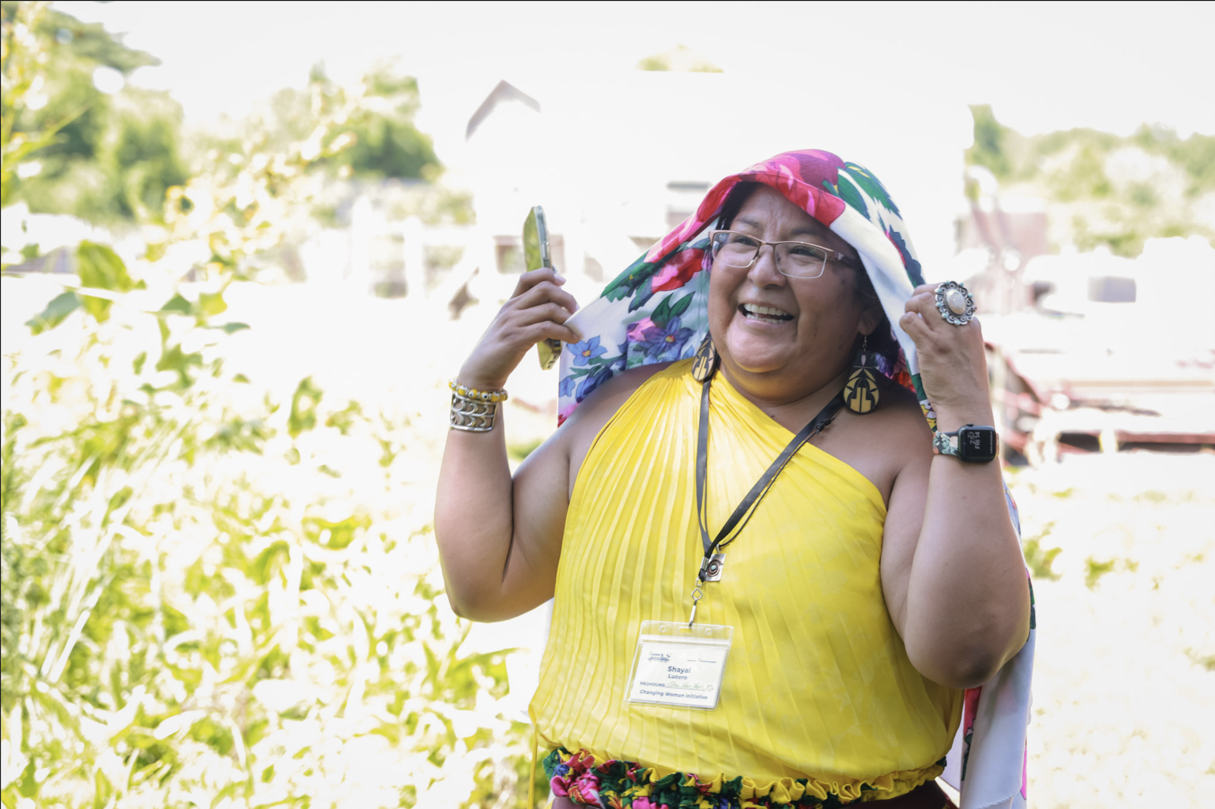 A woman wearing glasses and a yellow dress, smiling and adjusting a colorful headscarf outdoors with greenery and buildings in the background.