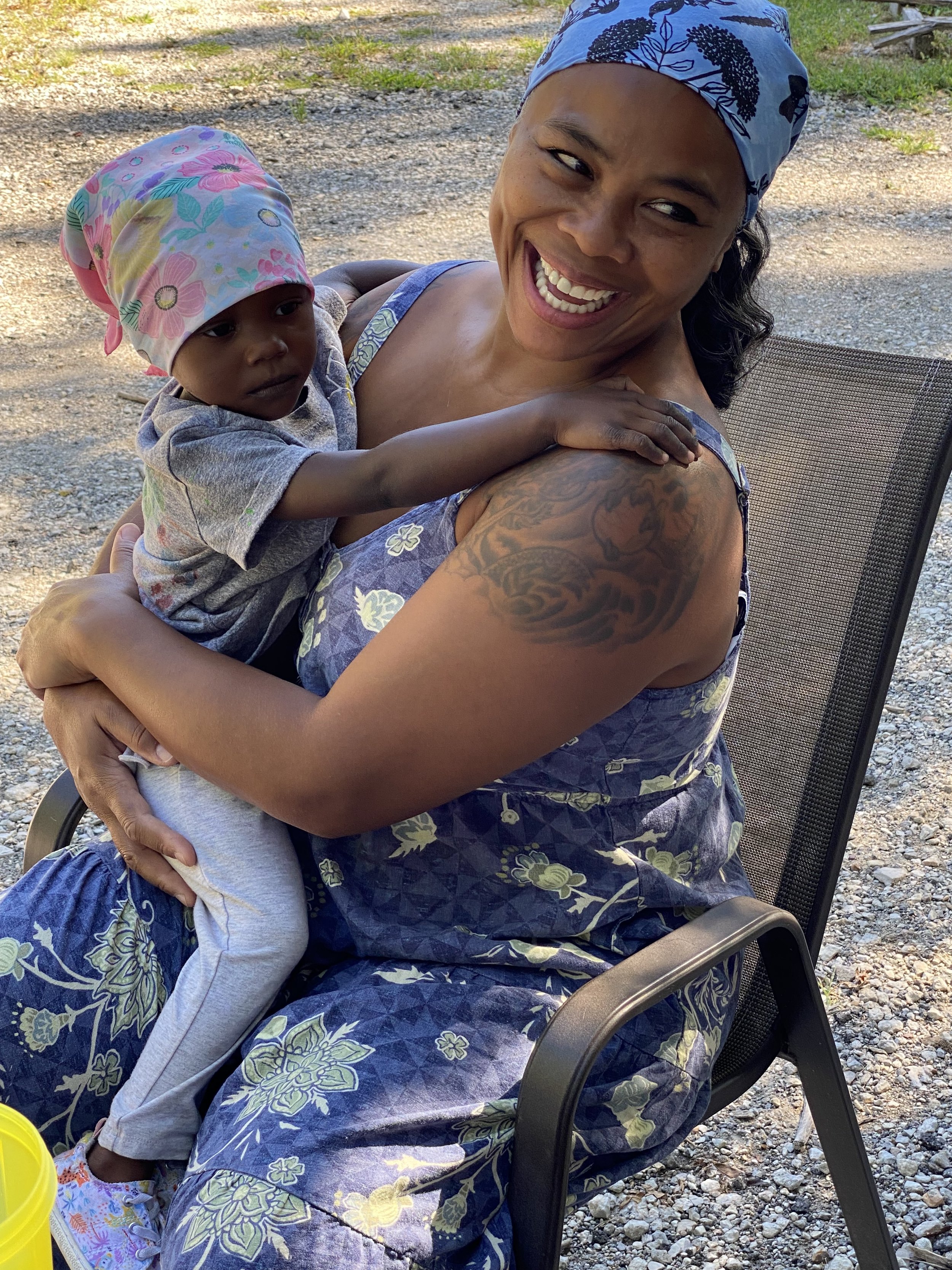 A woman with a blue bandana on her head and a tattoo on her arm is sitting outdoors, smiling while holding a young child who is resting on her shoulder.