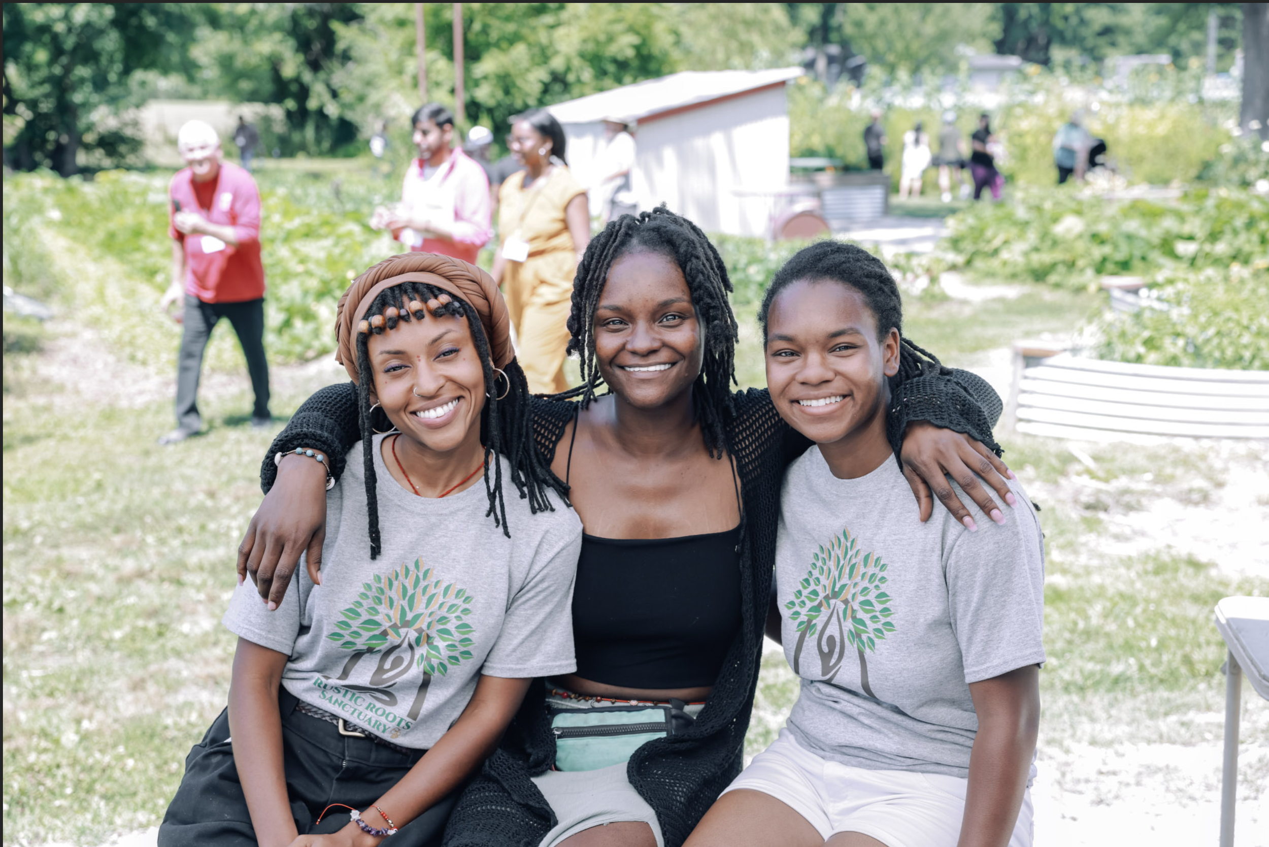 Three smiling women sitting outdoors with their arms around each other, at an outdoor community event or gathering.